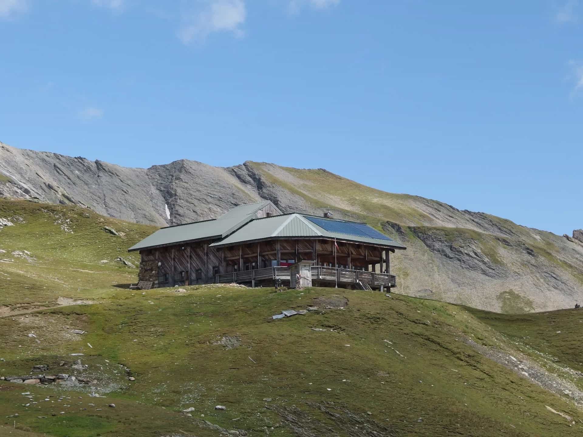 Wooden mountain refuge with solar panels on grassy slope below rocky peaks, Refuge de la Croix du Bonhomme.