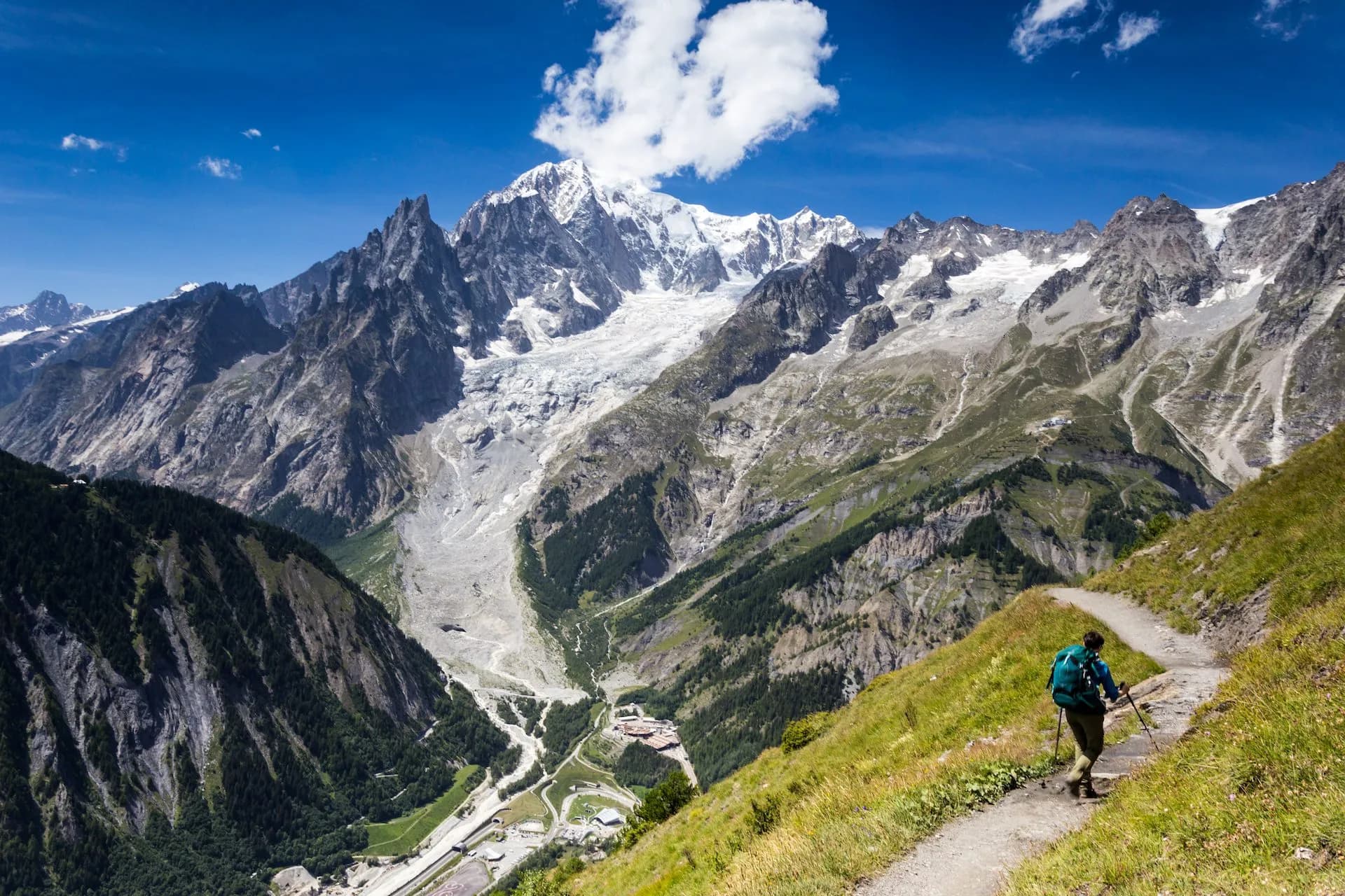 Hiker on dirt trail with snow-capped alpine mountains and glacier under blue sky