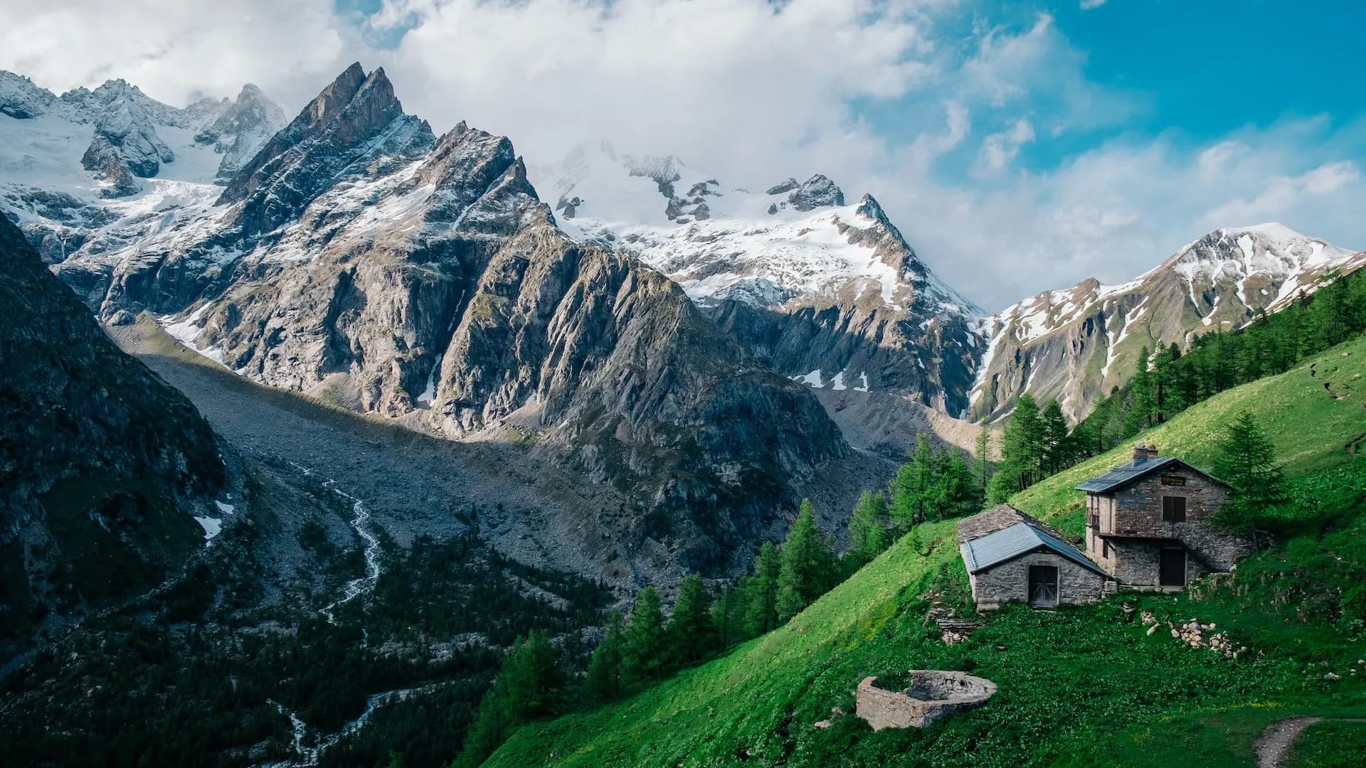 Stone refuge huts on green slope below snow-capped alpine mountains near Refuge Bonatti.