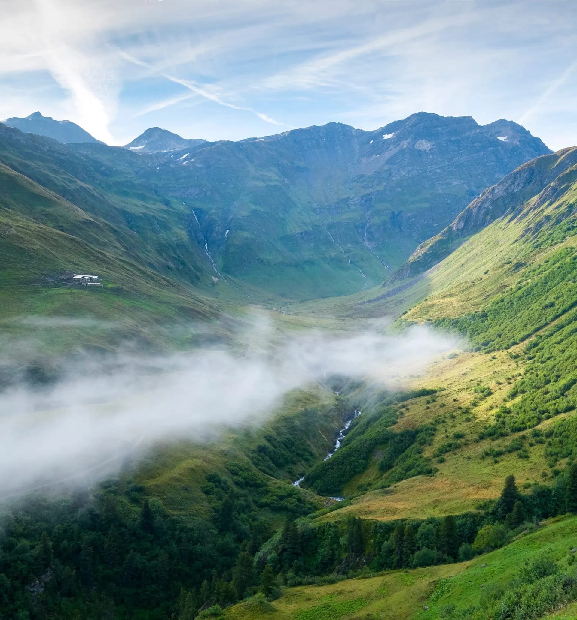 Mountain valley with green slopes, low fog, and a stream under a blue sky with contrails.