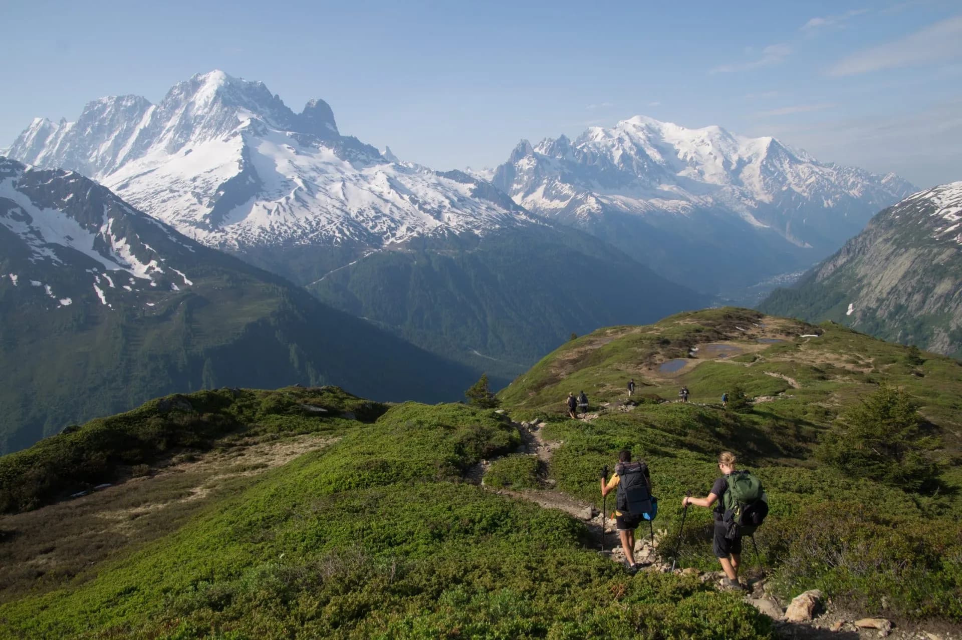 Hikers with backpacks trekking on a ridge overlooking snow-capped mountains and a deep valley.