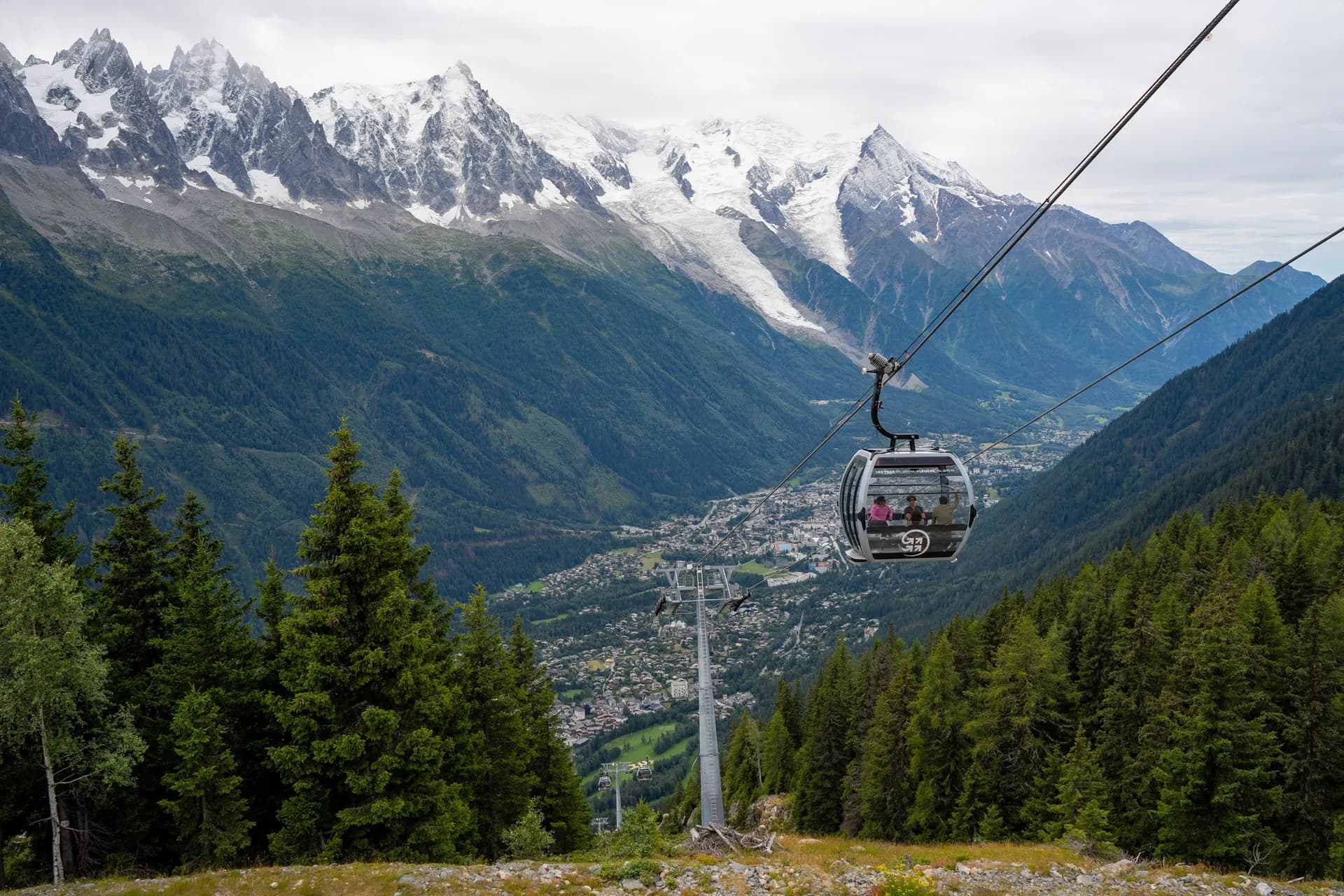 Cable car descending over forested slopes toward Chamonix valley with snow-capped mountains.