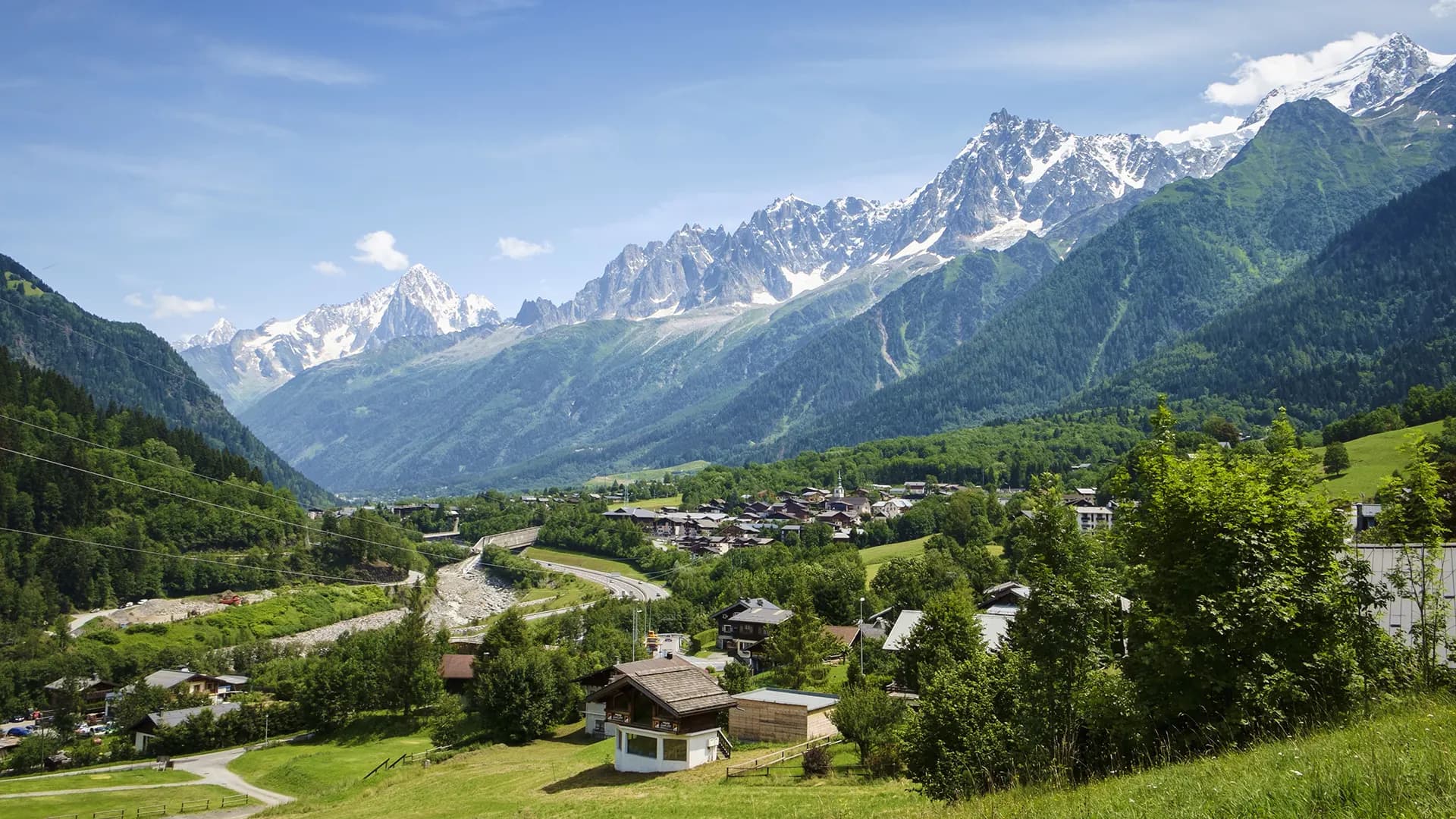 Alpine village nestled in a valley with snow-capped mountains under a blue sky.
