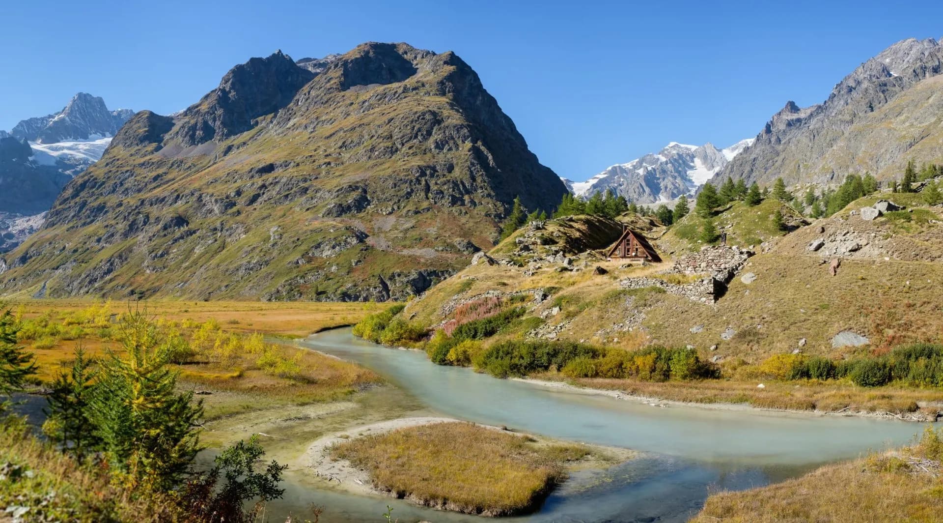 Mountain valley with winding river, small wooden cabin, and snow-capped peaks under blue sky.