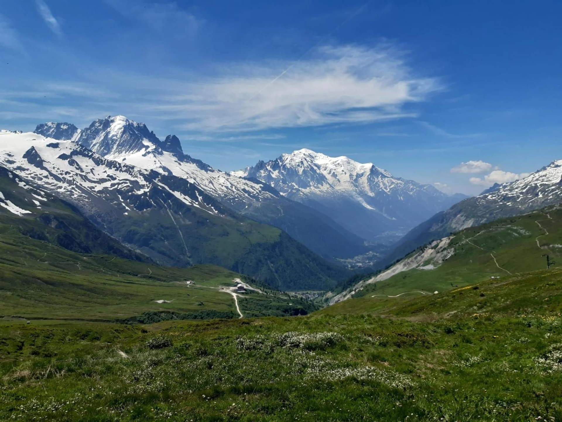 Alpine trail view with snow-capped mountains, green slopes, and a deep valley under a blue sky.