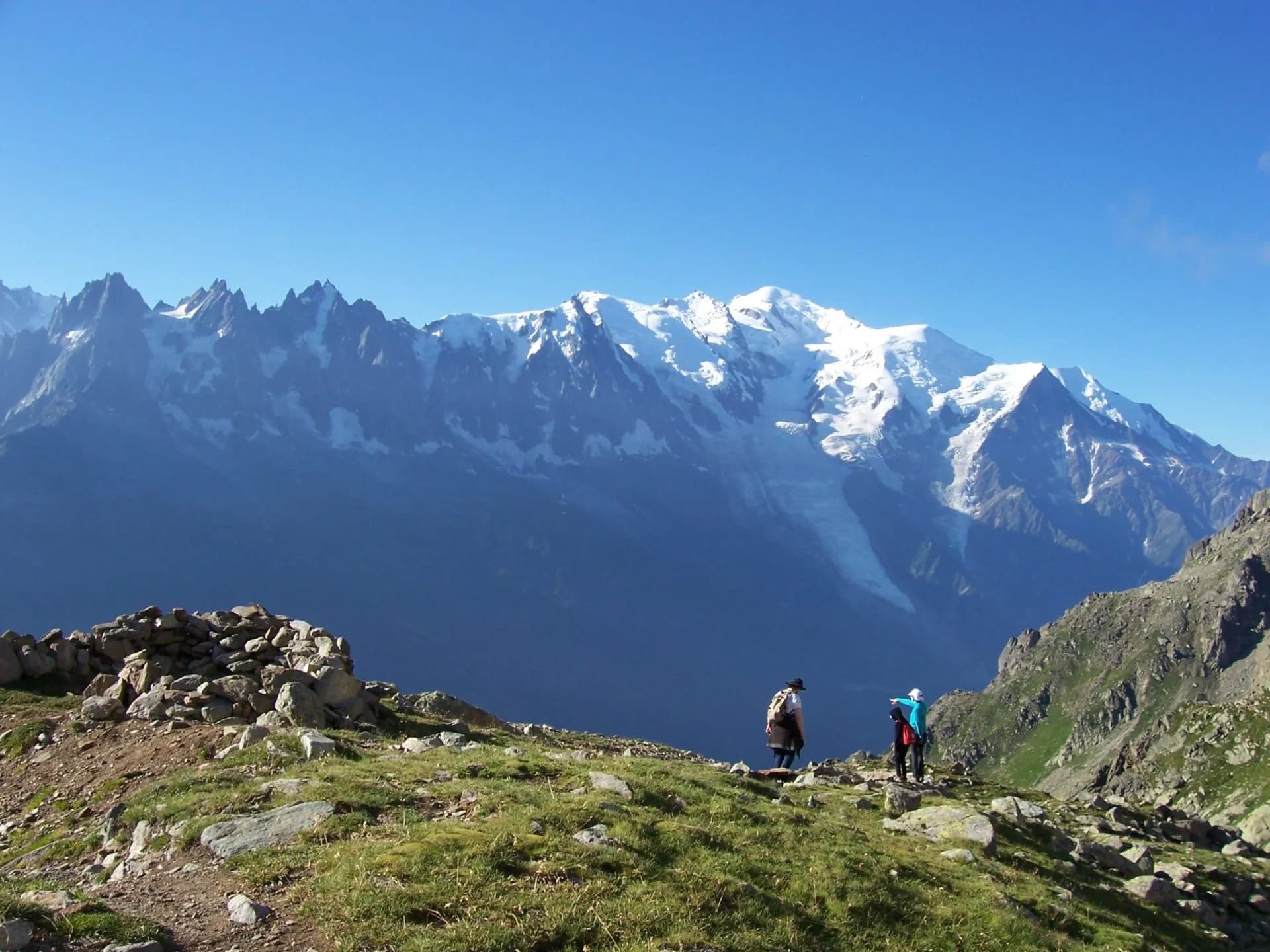 Hikers on grassy ridge overlooking snow-capped mountains, likely Le Brevent.