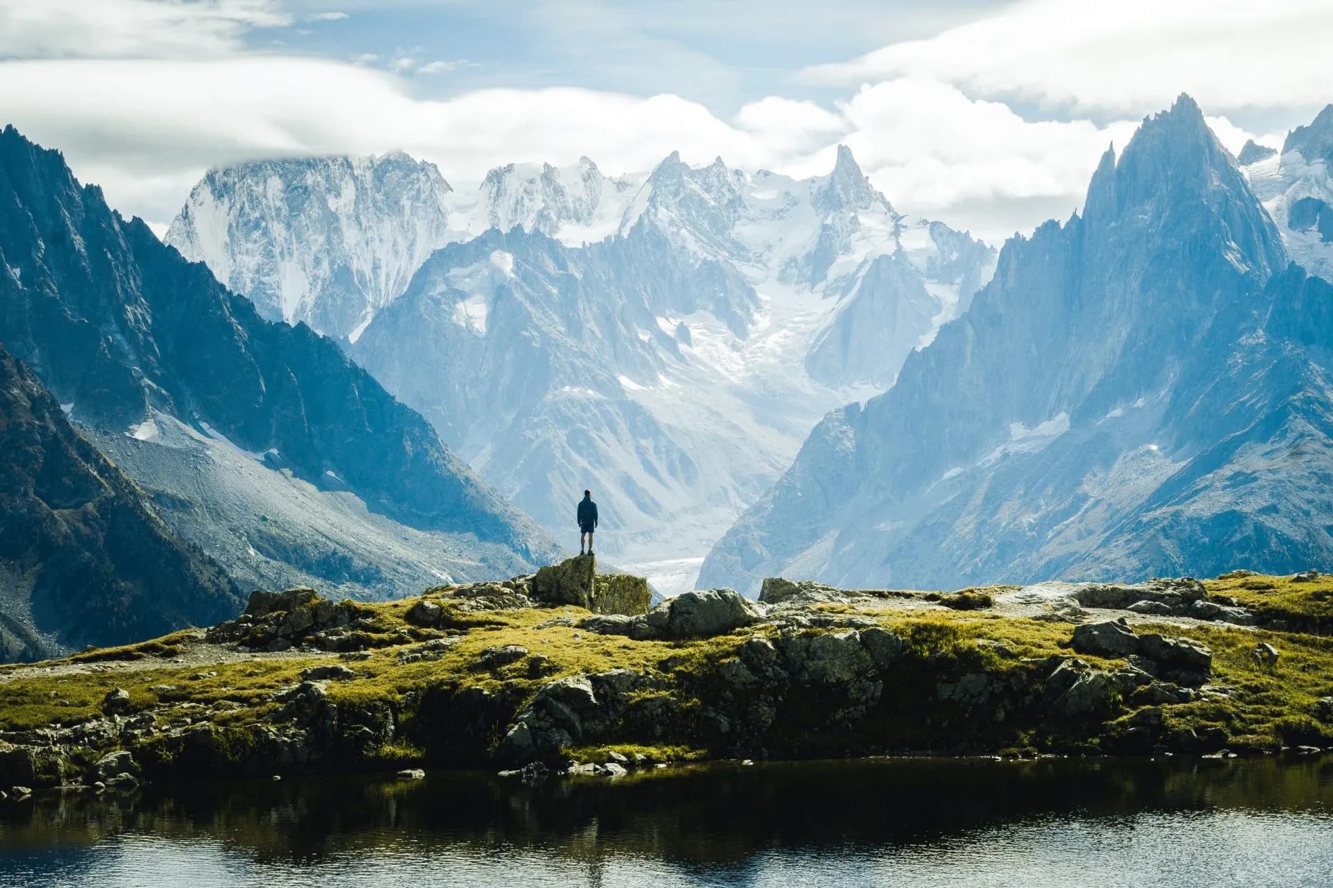 Hiker standing on rocky outcrop overlooking alpine lake with snow-capped mountains in background.