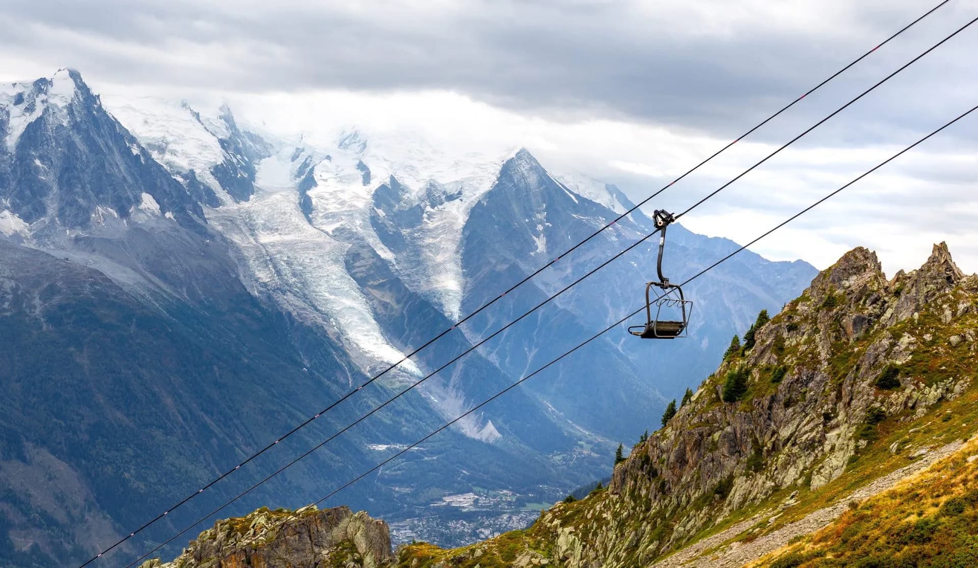 Empty chairlift ascending rocky slope with snow-capped mountains and glacier in background, La Flégère.