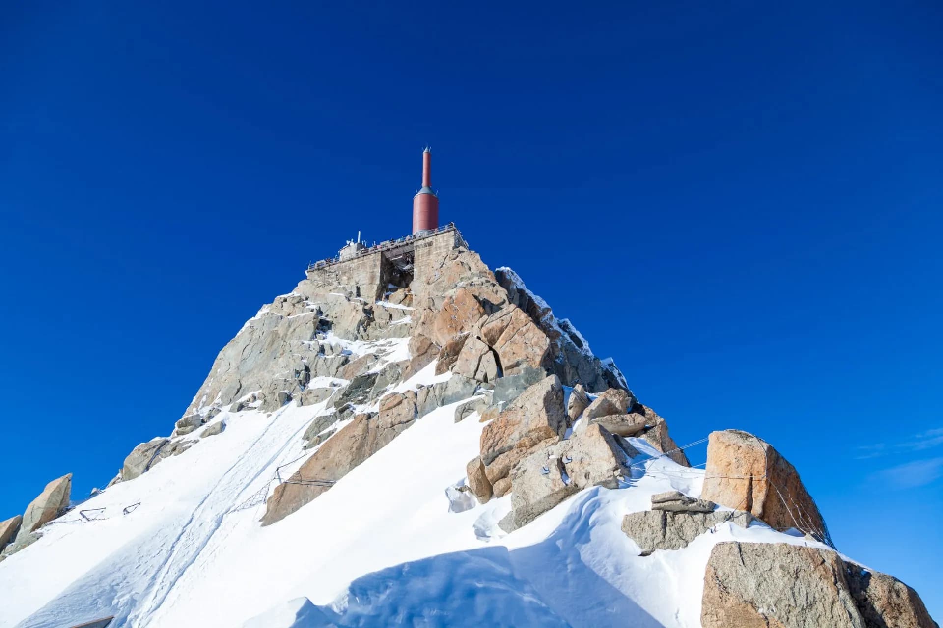 Aiguille du Midi summit station with red antenna on snow-covered granite peak under bright blue sky.