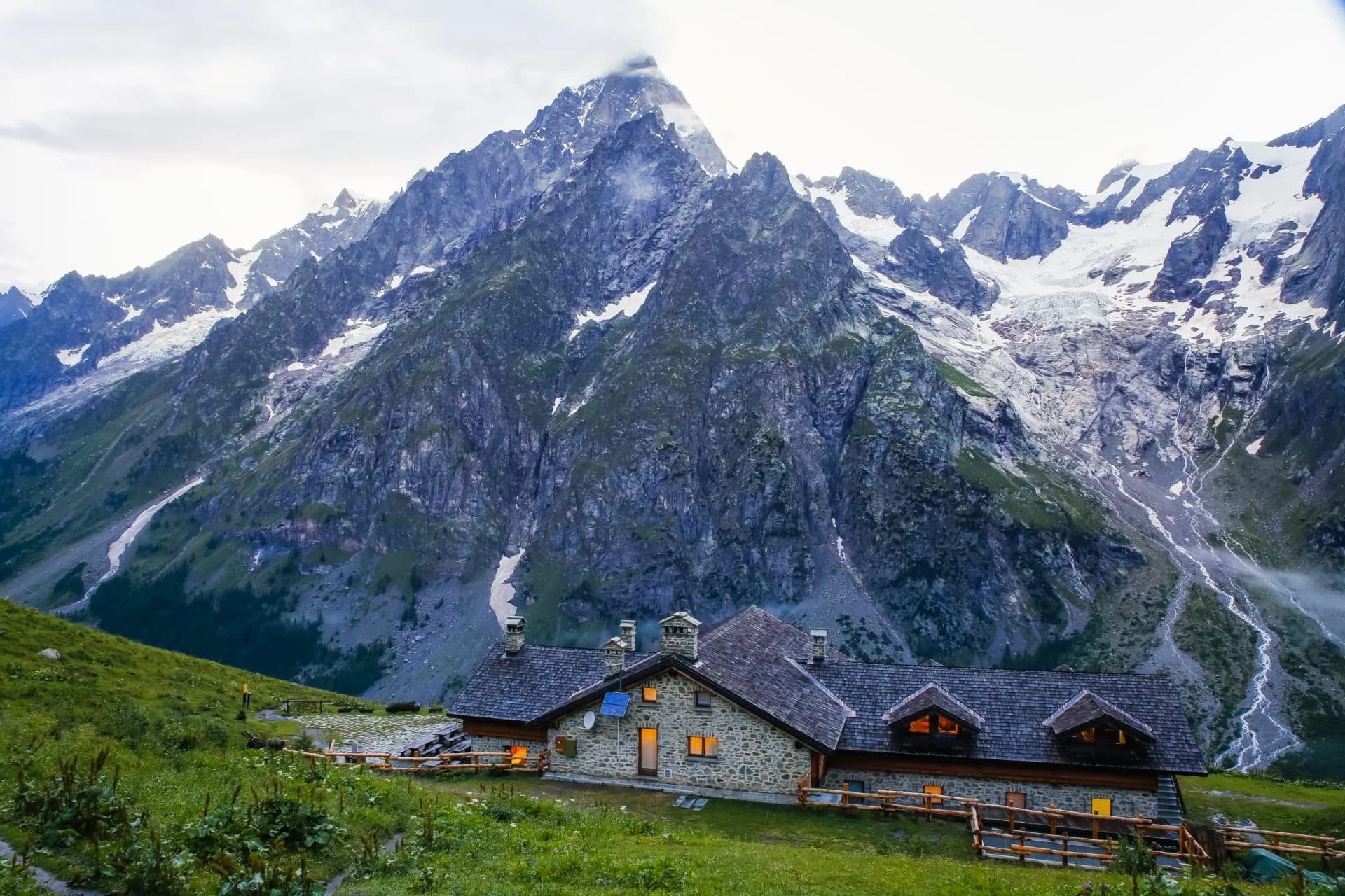 Rifugio Bonatti stone mountain hut nestled in green alpine meadow below rocky, snow-capped peaks.