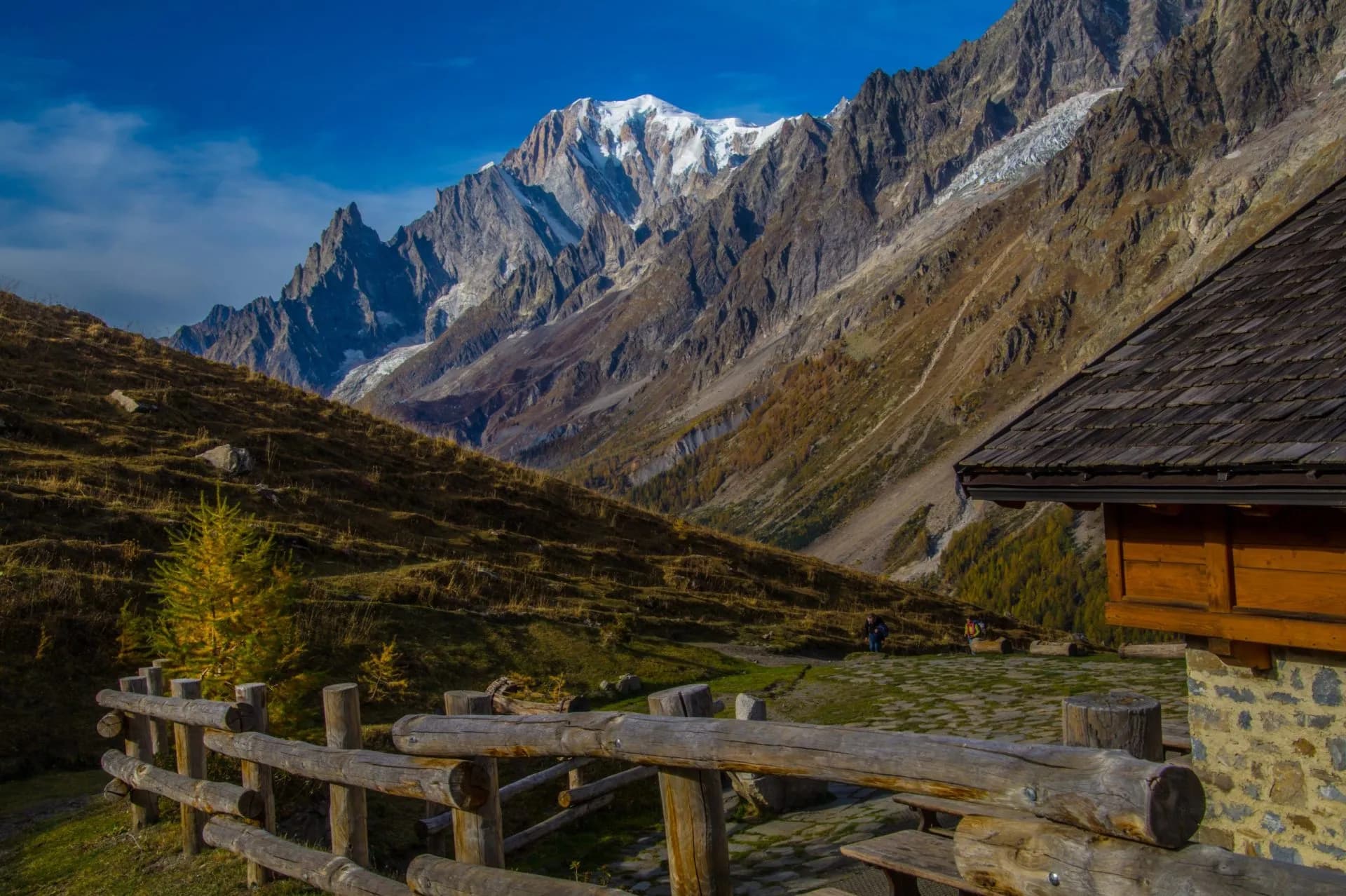 Rifugio Bonatti with snow-capped alpine mountains and autumn foliage under a blue sky.