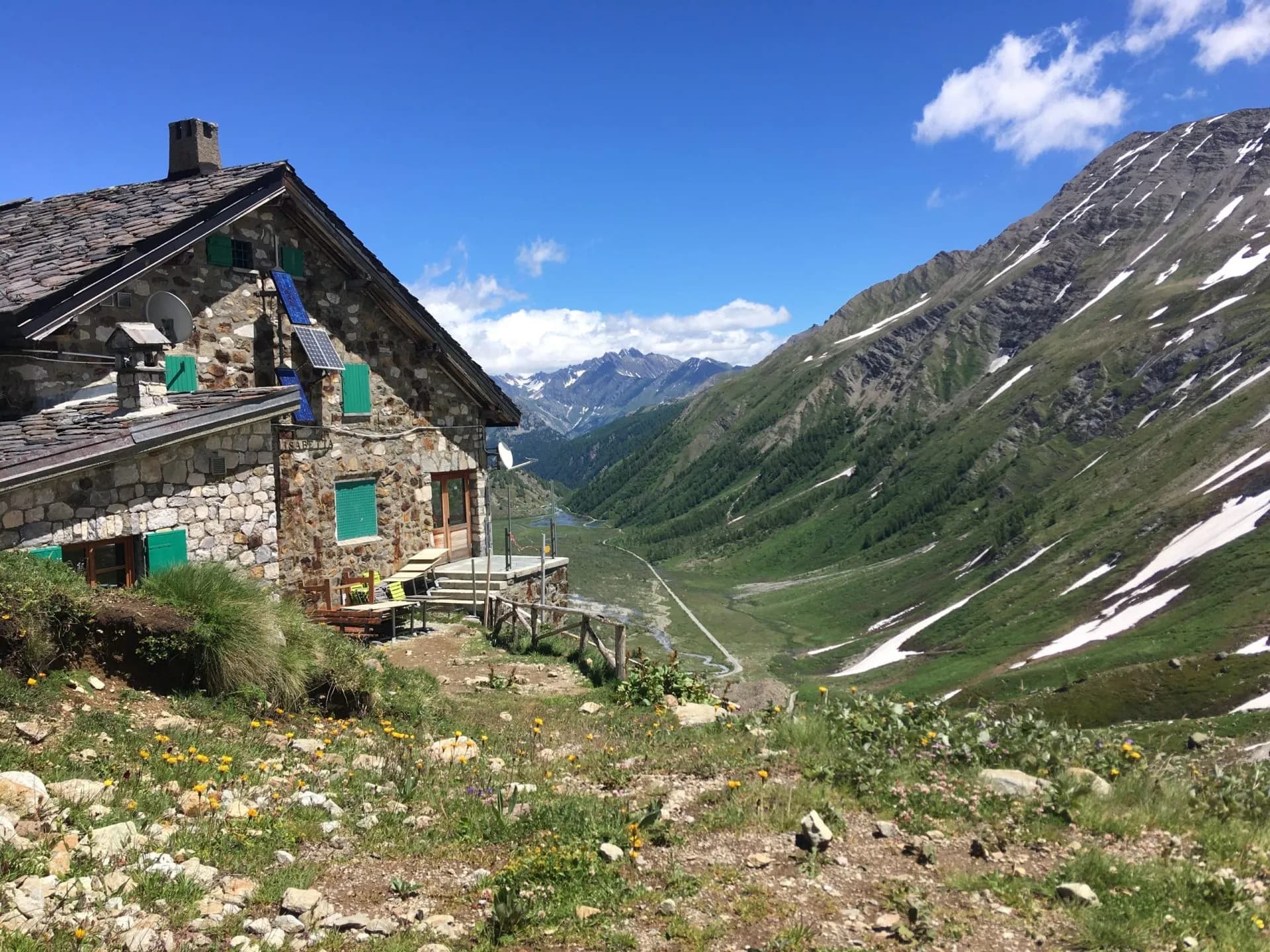 Rifugio Elisabetta stone mountain hut overlooking a green valley with snow patches under a blue sky.