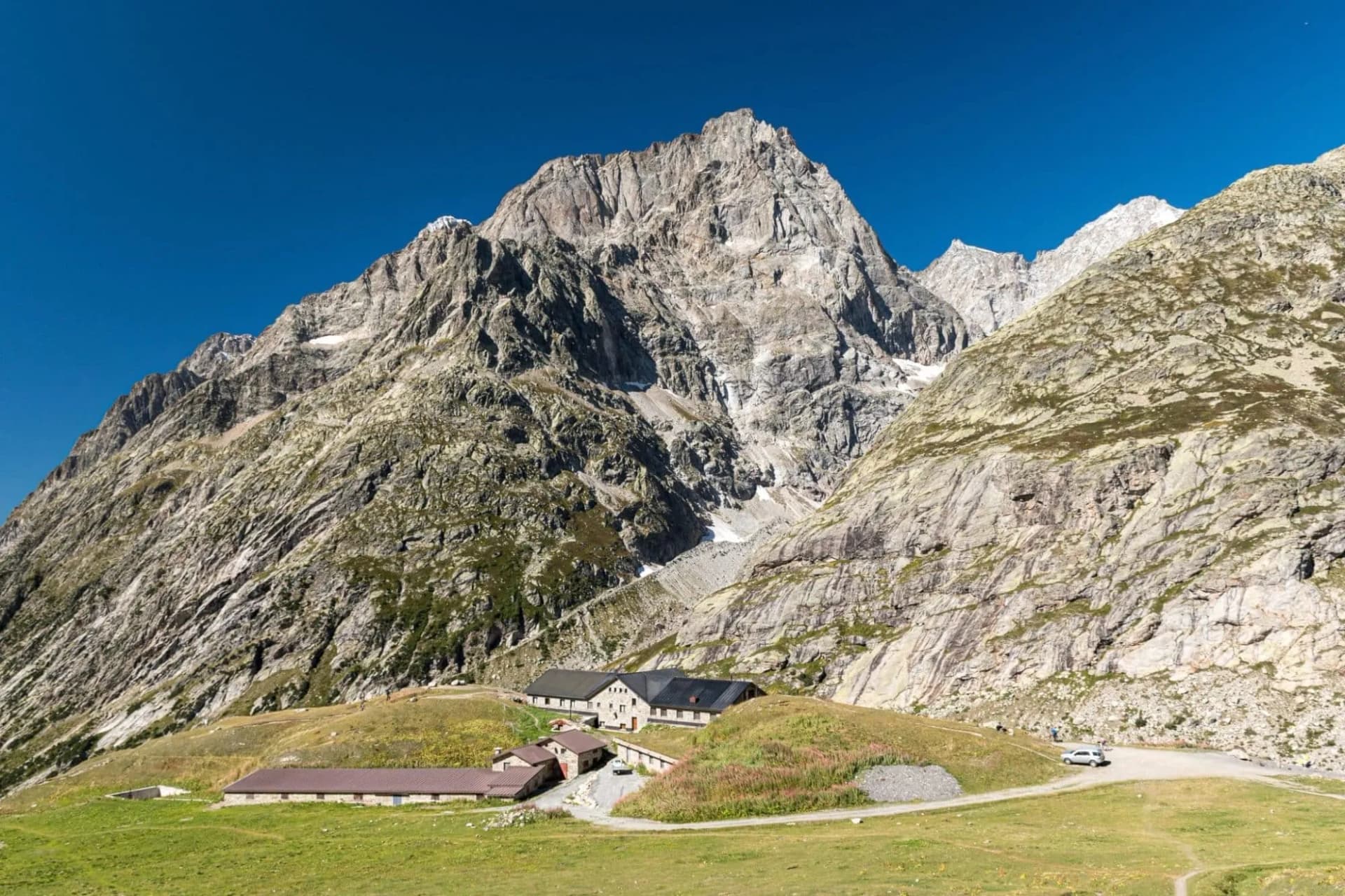 Mountain refuge buildings nestled below a massive, rugged peak under a clear blue sky.