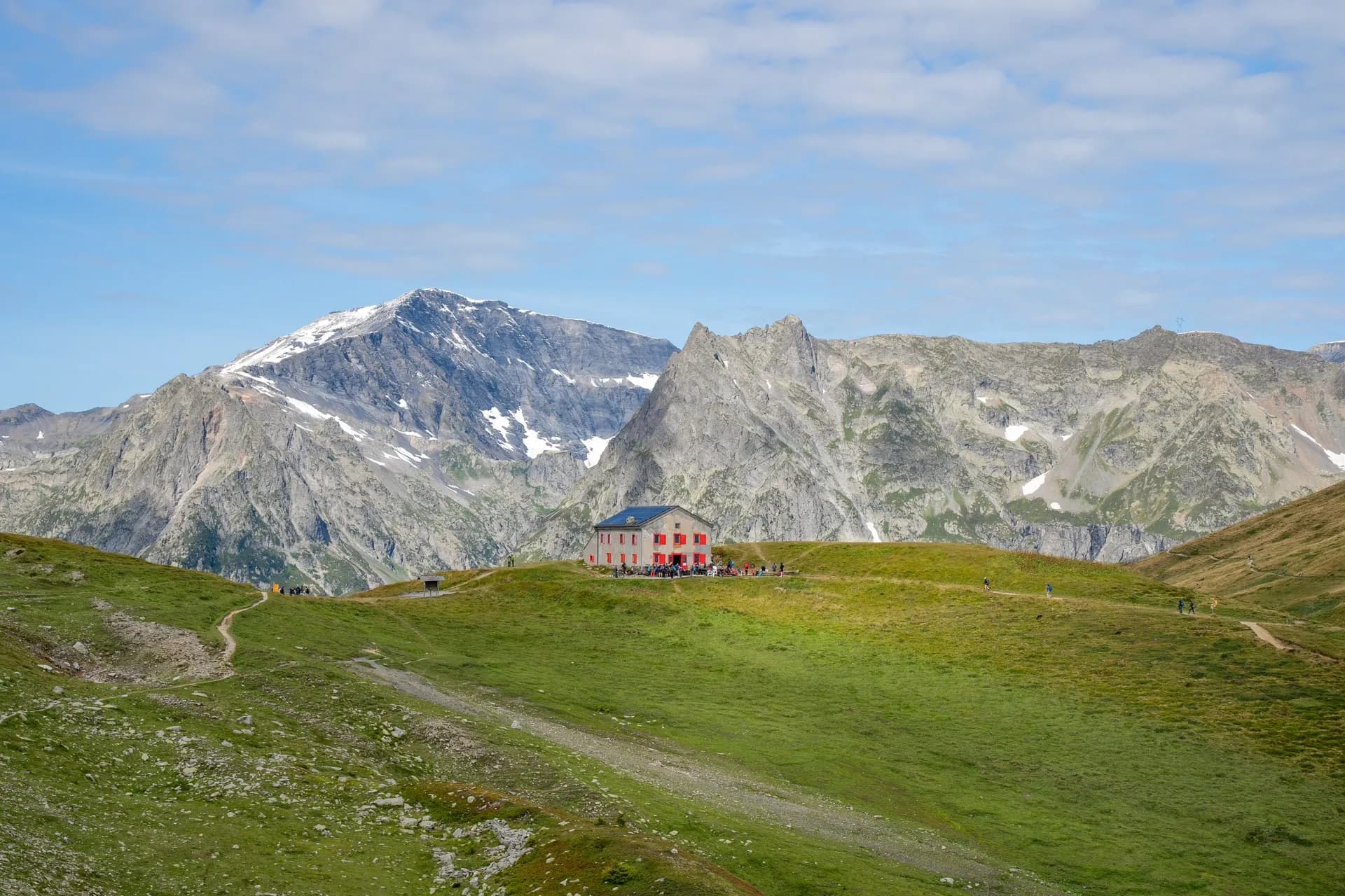 Refuge Col de la Balme with hikers on green alpine meadow below snow-capped mountains.