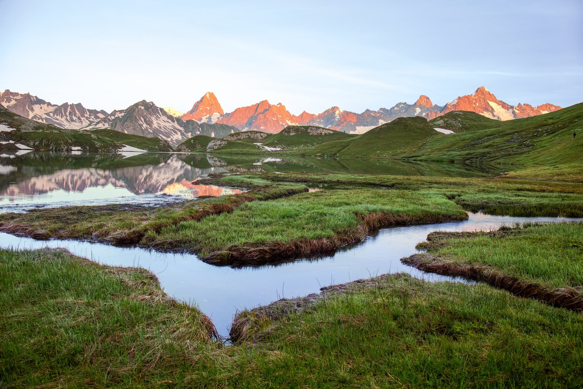 Grand Col Ferret wetlands with green grass, stream, and snow-capped mountains reflecting at sunrise.