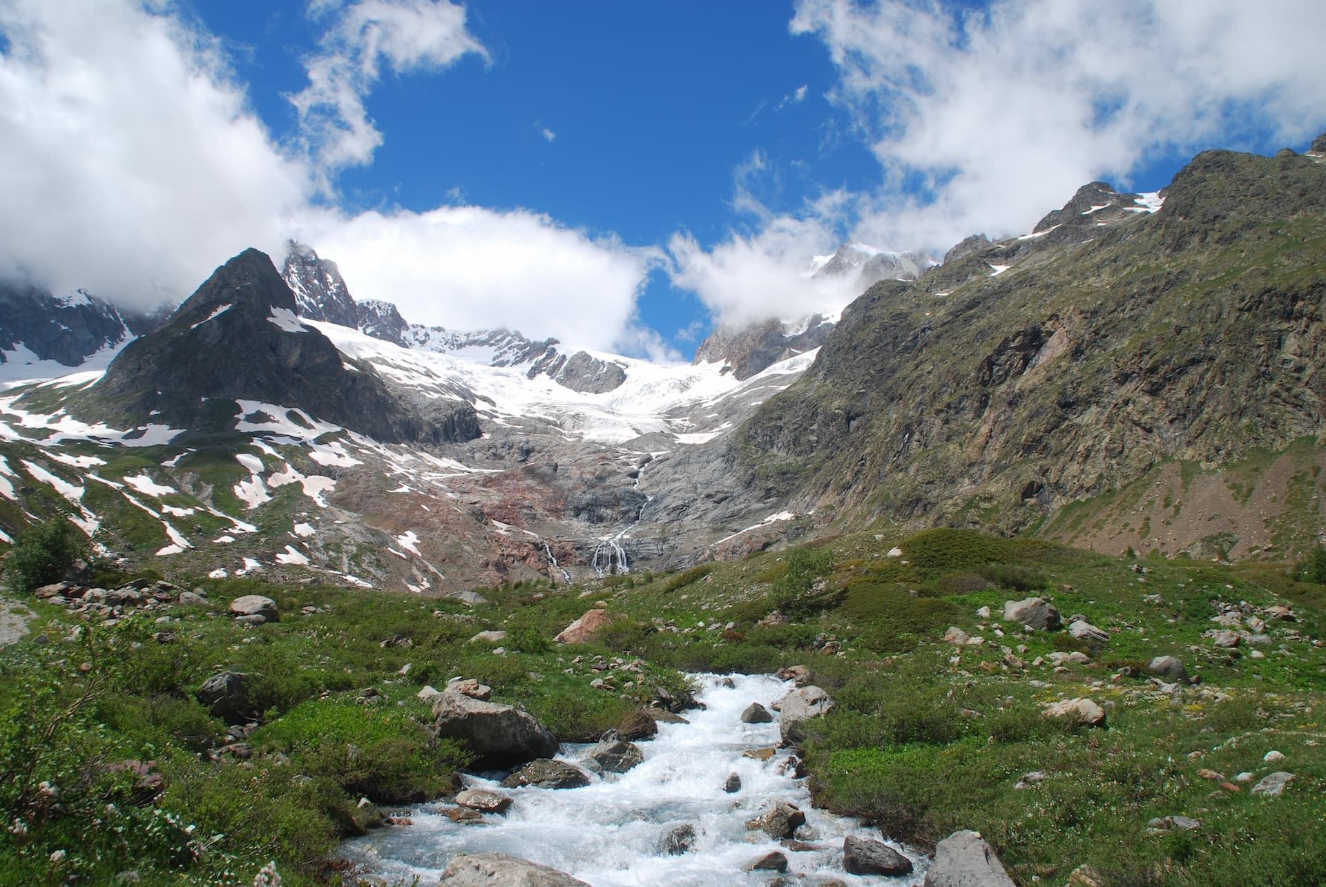 Mountain stream flowing from glacier under blue sky with clouds near Col de la Seigne.