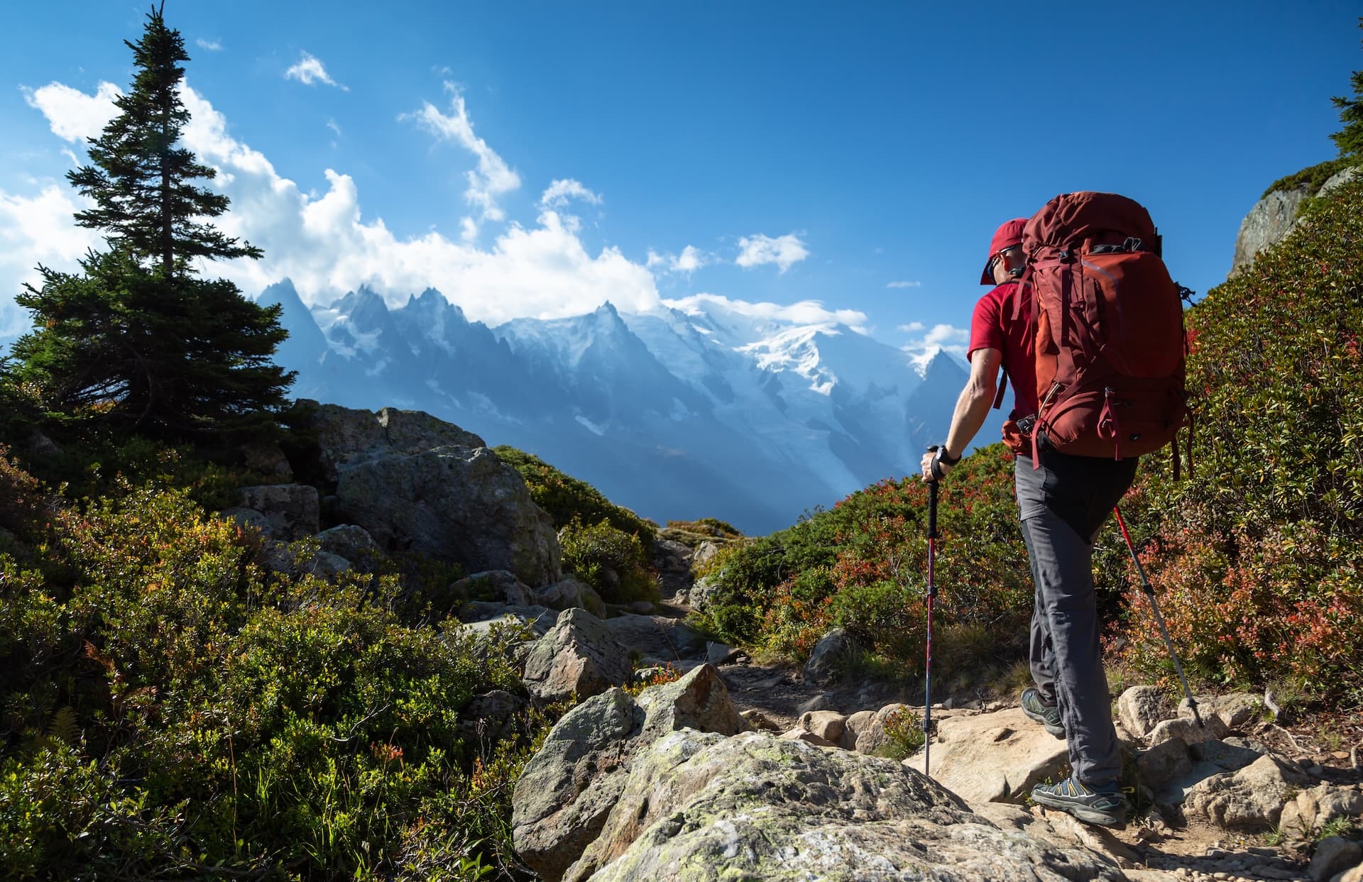 Hiker with large backpack hiking on rocky trail toward snow-capped mountains under blue sky.