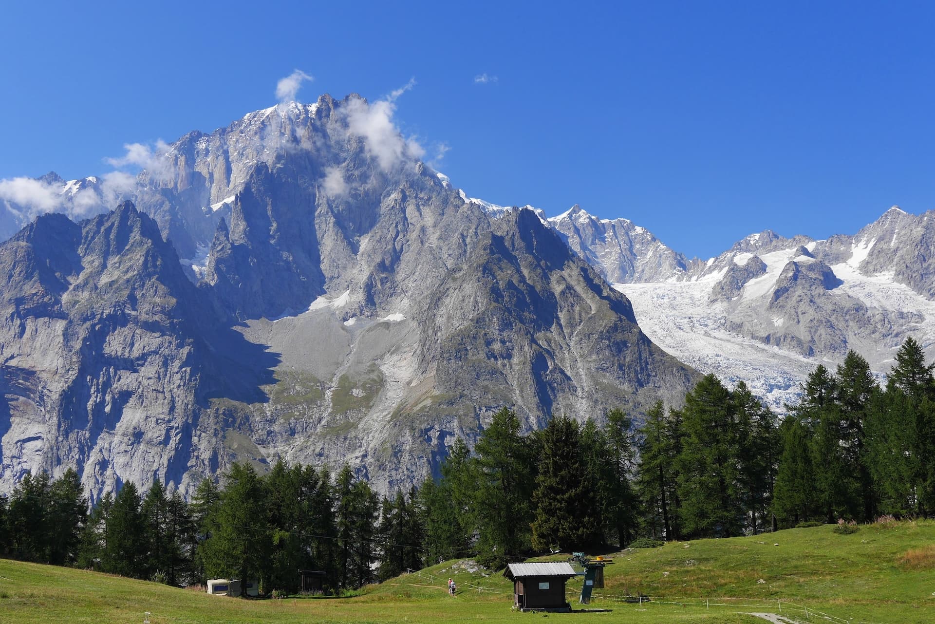 Scenic meadow with small hut, pine trees, and massive snow-capped mountains under Col de la Seigne.