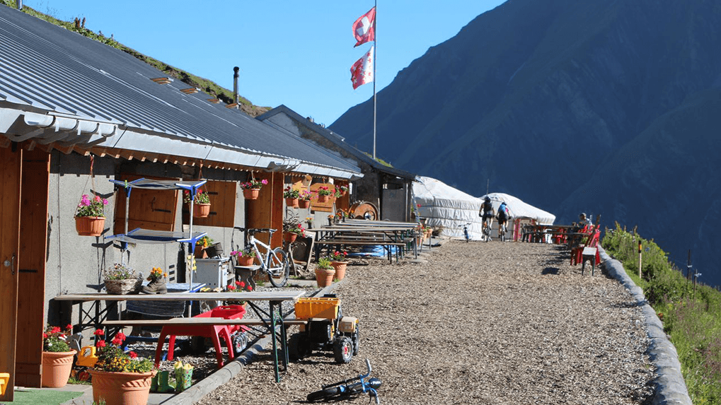 Mountain refuge with flower pots and picnic tables, cyclists near yurts below steep mountains.