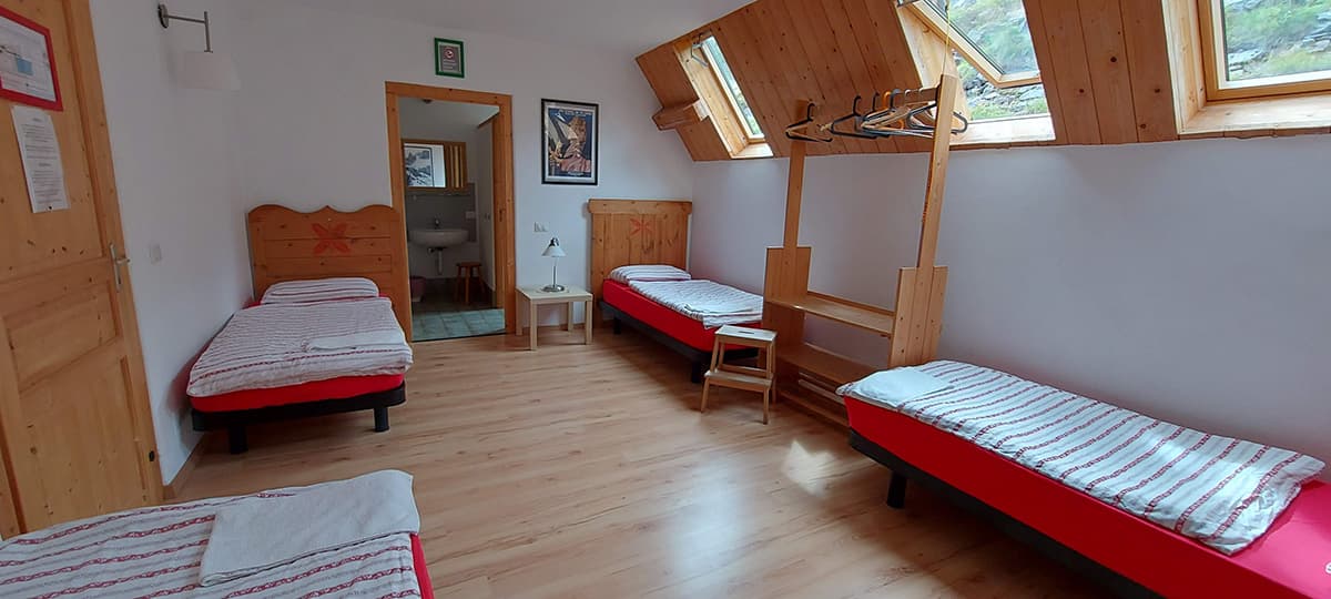 Dormitory room with four single beds, wood floors, and slanted windows showing mountain view.