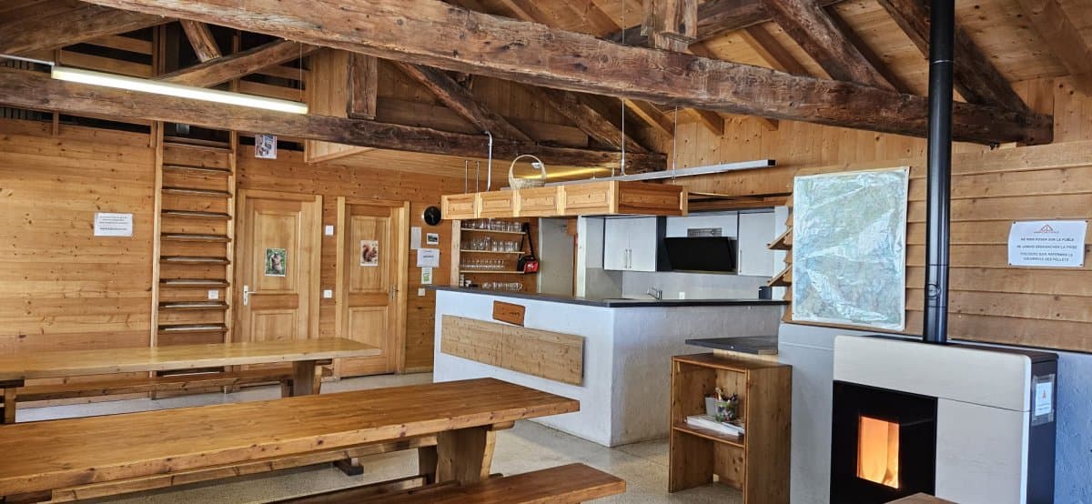 Interior of a rustic wooden mountain refuge dining area with long tables and a small kitchen counter.