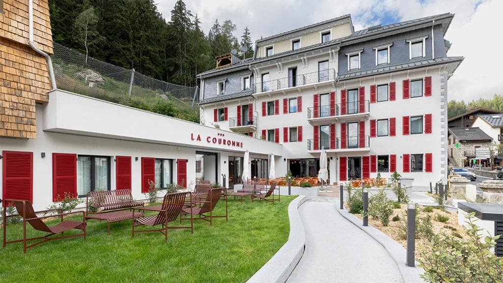 Hotel La Couronne exterior with white facade, red shutters, and lawn seating near forested mountain slope.