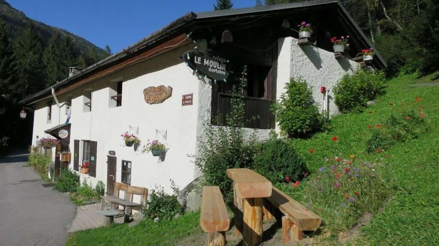 White stone building, "Le Moulin d'Artoise," with wooden benches on a grassy slope near a mountain road.
