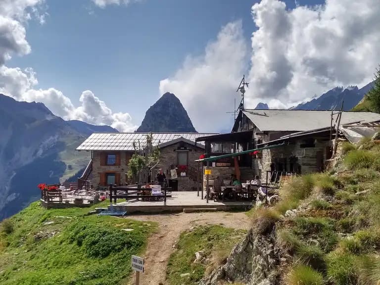 Rifugio Bertone mountain hut with outdoor dining terrace set against dramatic alpine peaks under a cloudy sky.