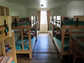 Dormitory room with multiple wooden bunk beds, green mattresses, and a window with curtains.