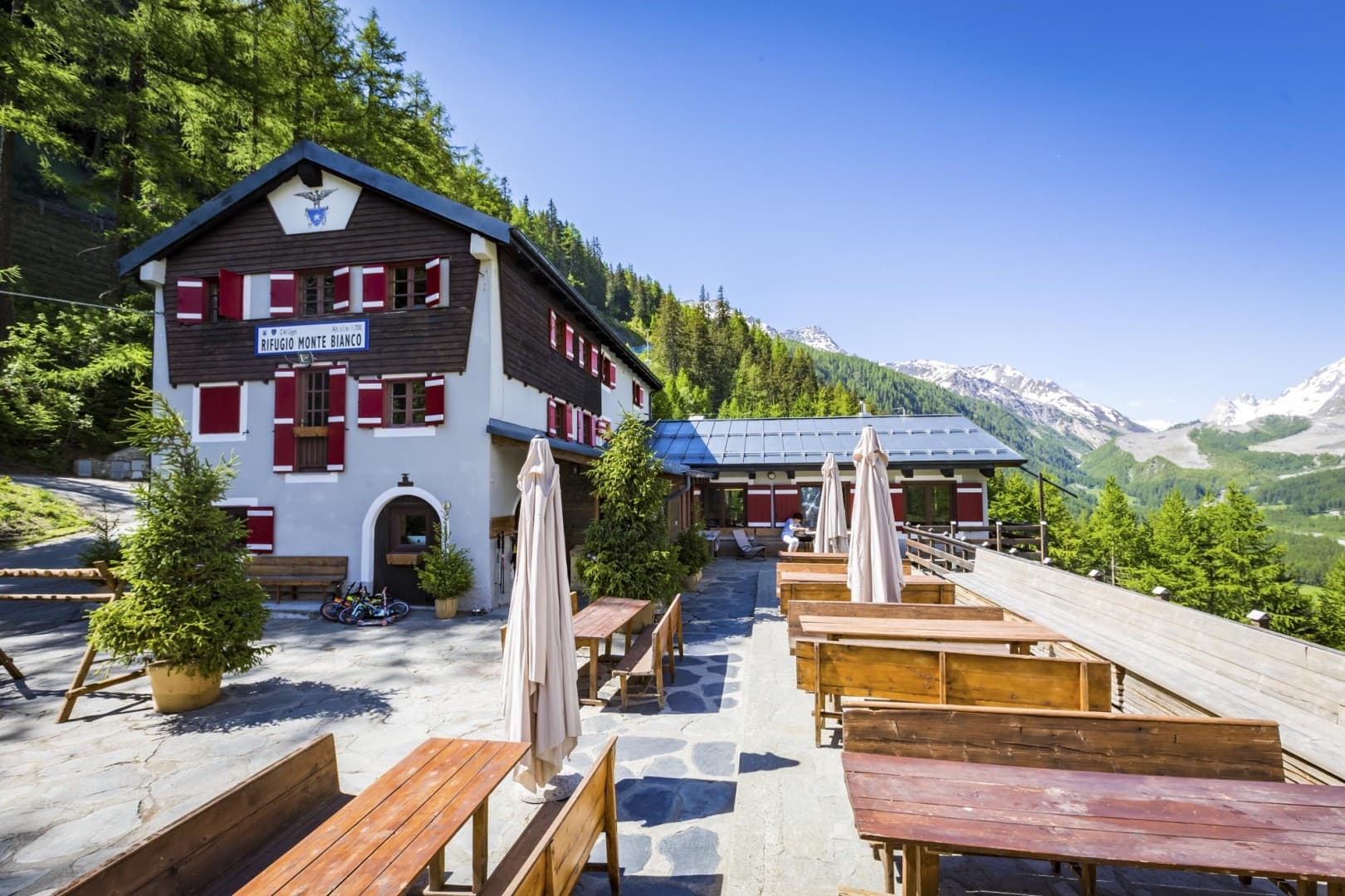 Rifugio Monte Bianco with outdoor wooden tables set against a backdrop of green forest and snowy mountains.