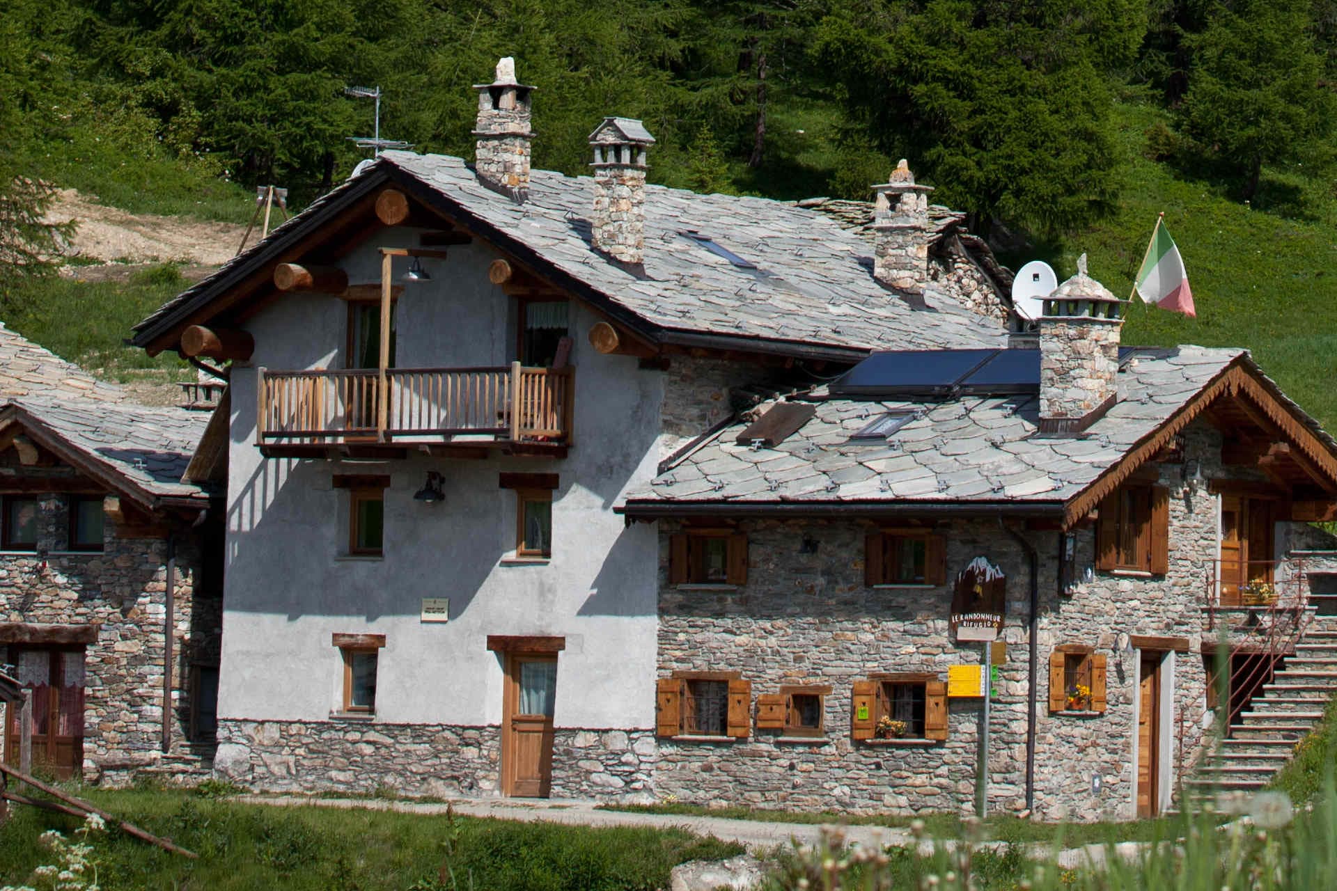 Stone refuge Le Randonneur with slate roof and Italian flag, set against a green mountain slope.