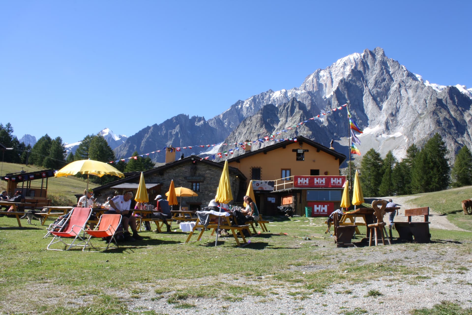 Rifugio Maison Vieille outdoor dining area with snow-capped mountains under clear blue sky.