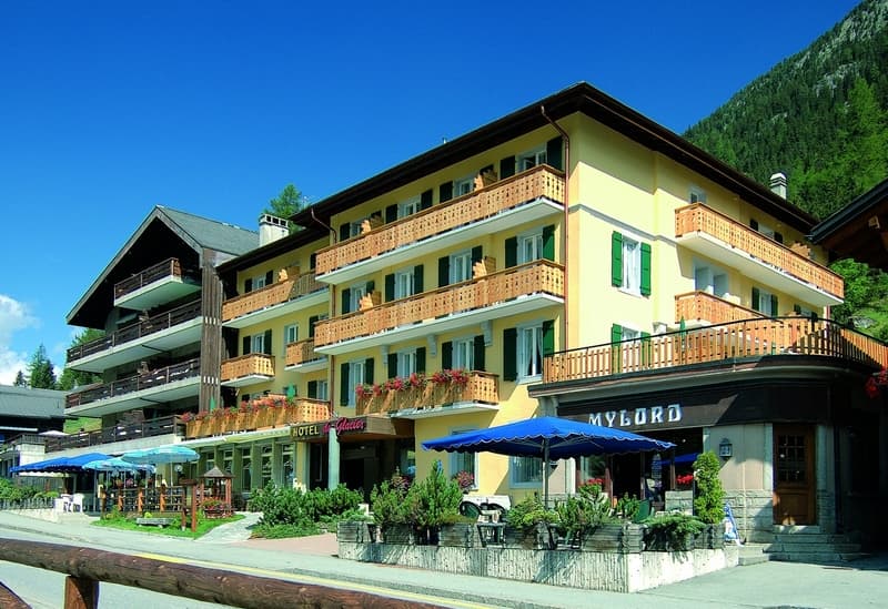 Hotel du Glacier building with wooden balconies and outdoor cafe seating against a forested mountain.