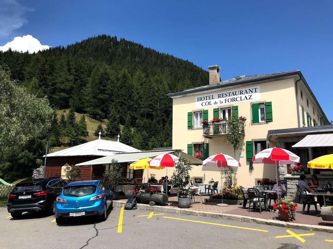 Hotel Restaurant Col de la Forclaz with outdoor dining, parked cars, and forested mountain backdrop.