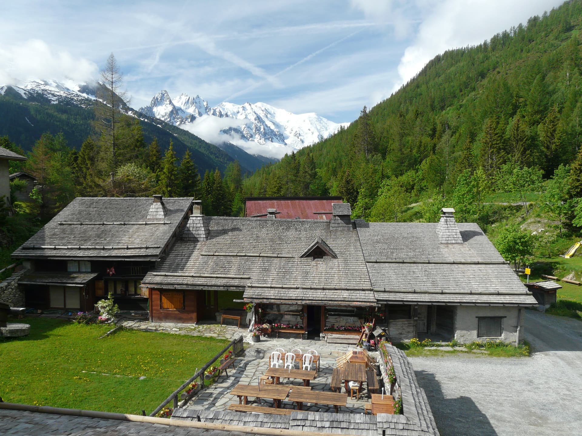 Chalet with wooden roof in mountains, outdoor dining area, and snowy peaks visible.