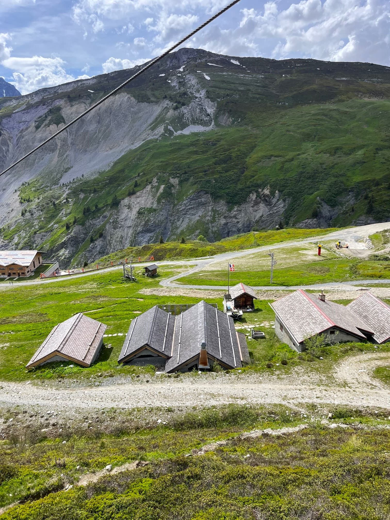 Mountain chalets near a ski lift cable on a green slope under a cloudy sky.