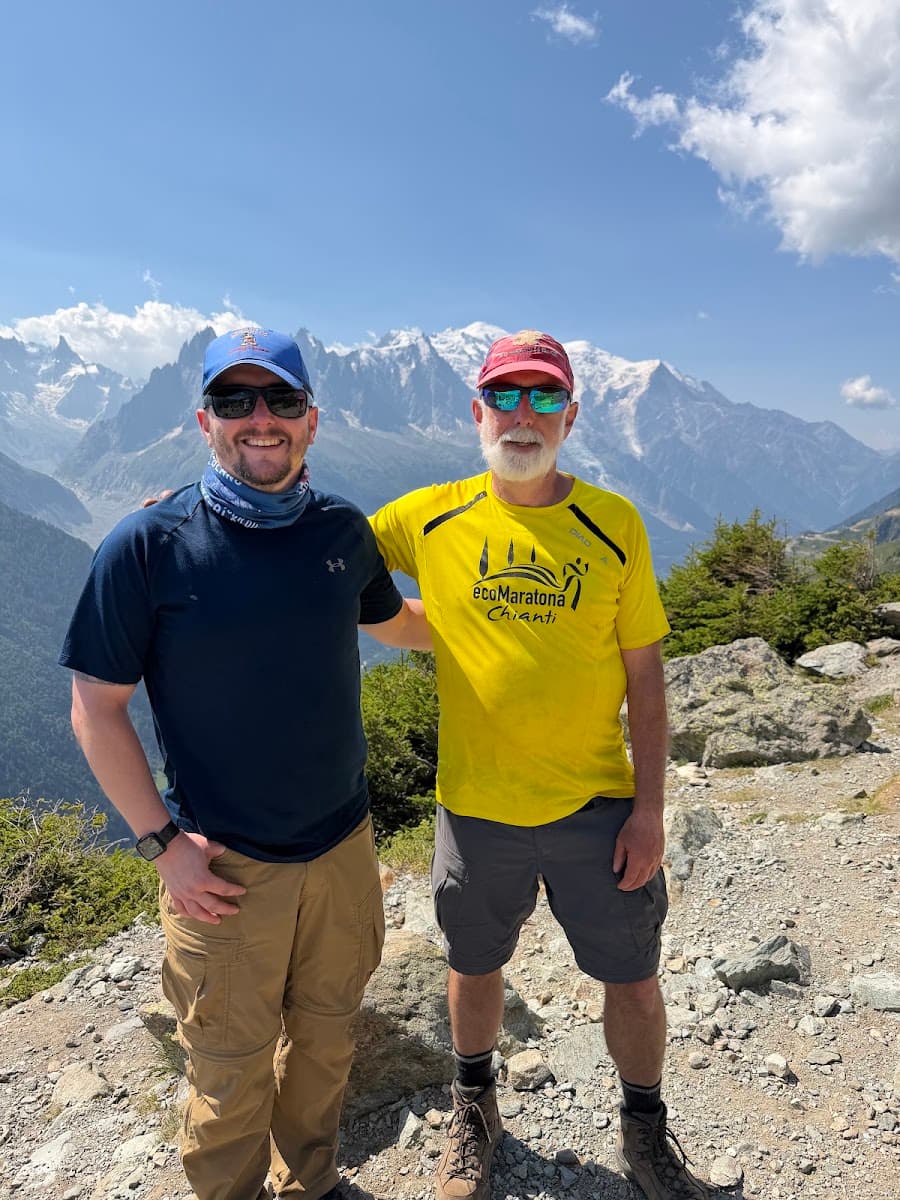 Two hikers standing on a mountain trail with snow-capped peaks in the background