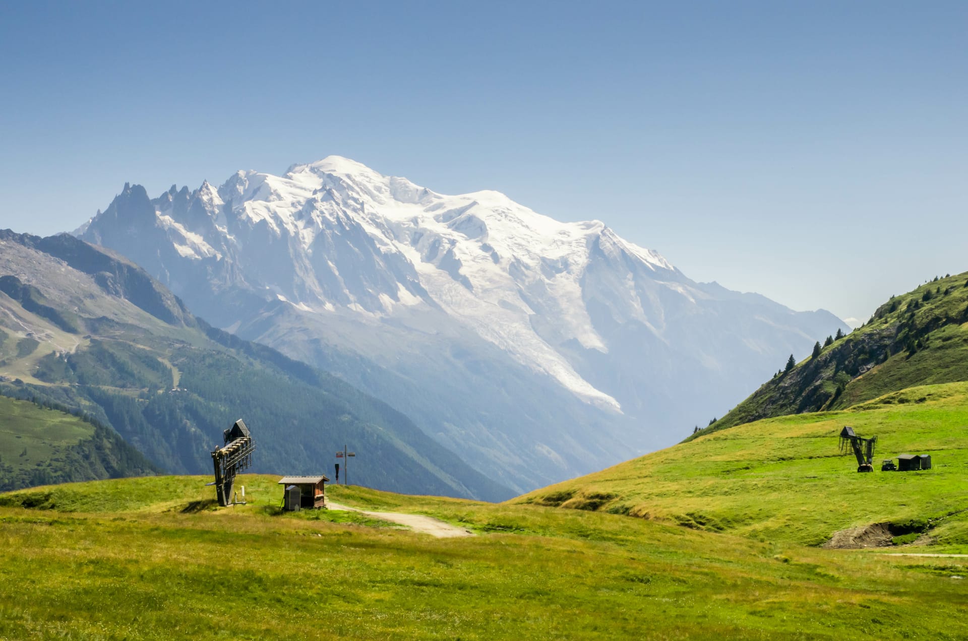 Snow-capped Mont Blanc massif towering over green alpine meadow with ski lift structures.