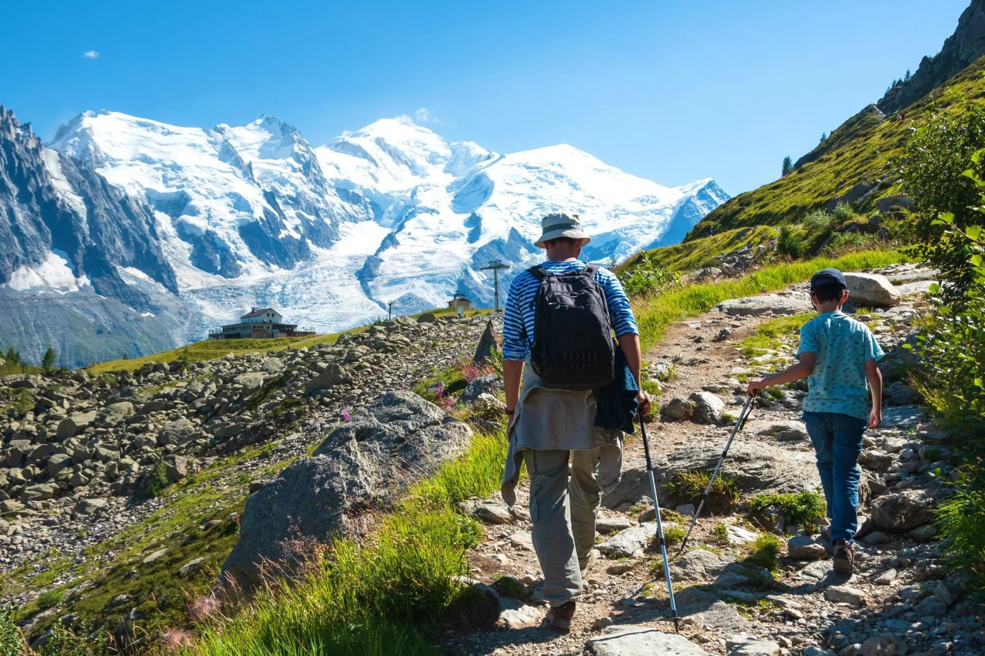 Hikers on rocky trail toward snow-capped mountains near Planpraz on a sunny day.