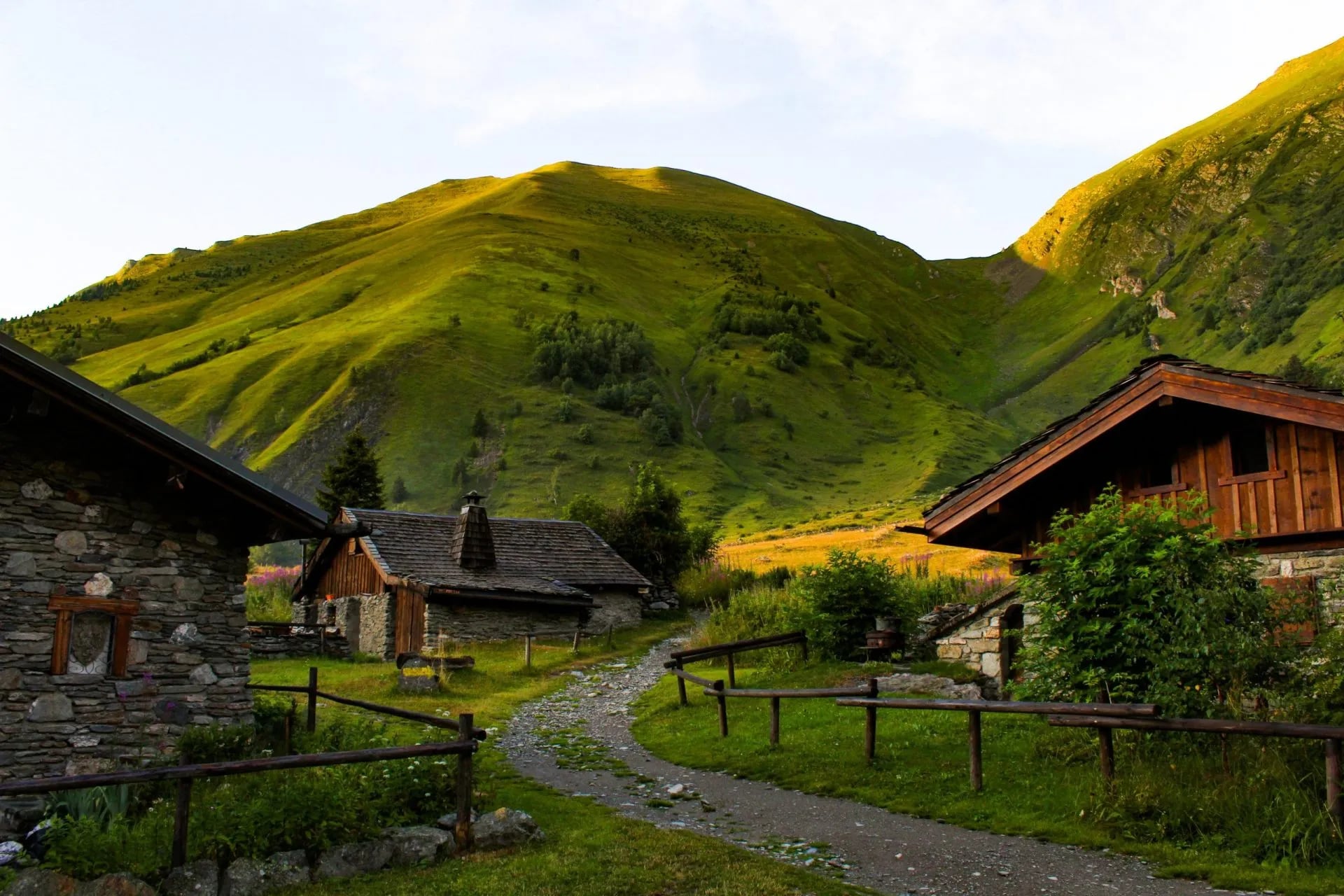Stone and wood alpine chalets near a gravel path below steep, grassy mountainsides.