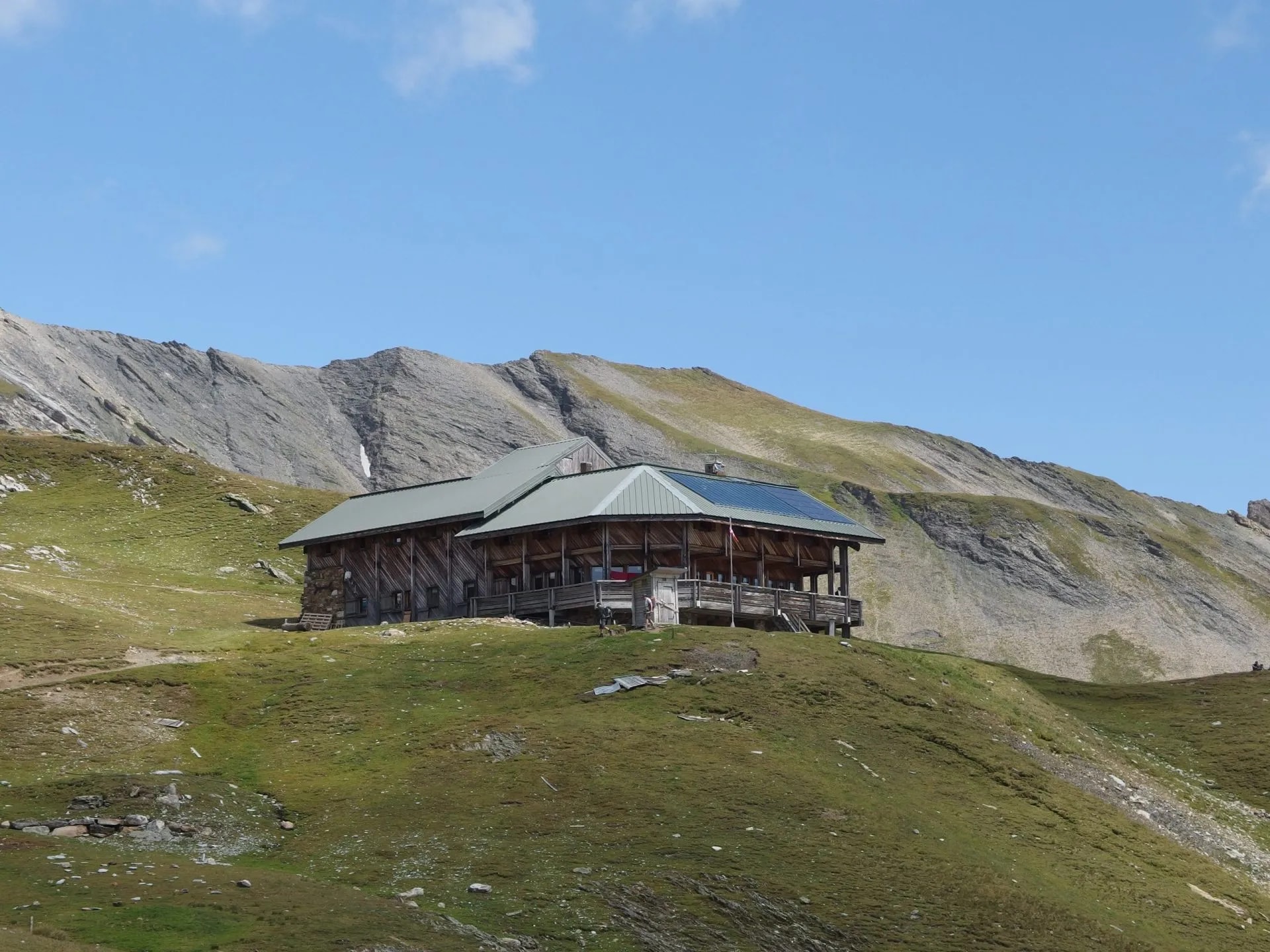 Wooden mountain hut with solar panels on grassy slope below rocky peaks under blue sky.