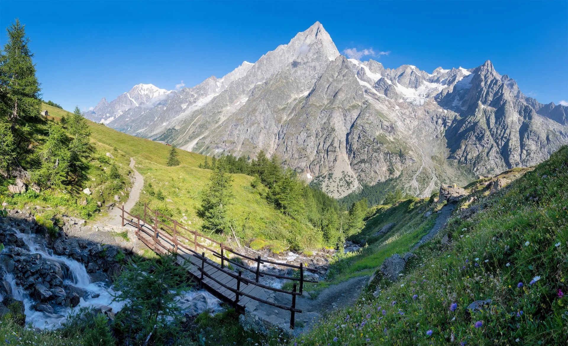 Hiking trail with wooden bridge over stream below the Grandes Jorasses massif in Val Ferret.