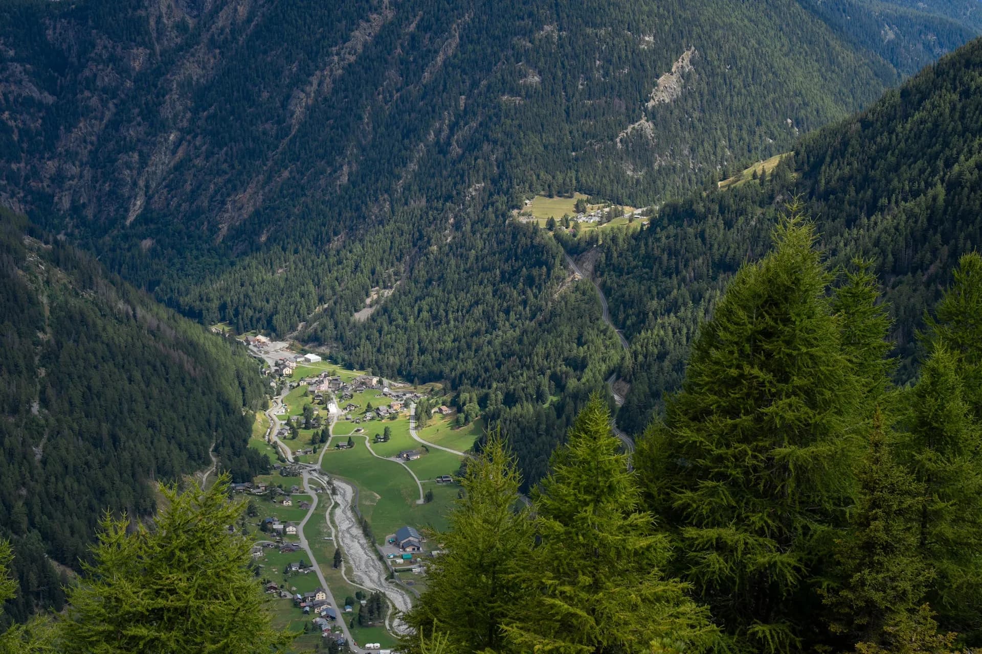 Alpine village nestled in a steep, forested valley with a riverbed, viewed from above.