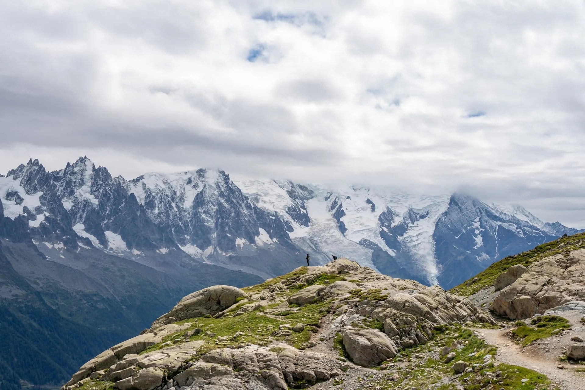 Hikers on rocky outcrop with massive snow-capped mountains under cloudy sky