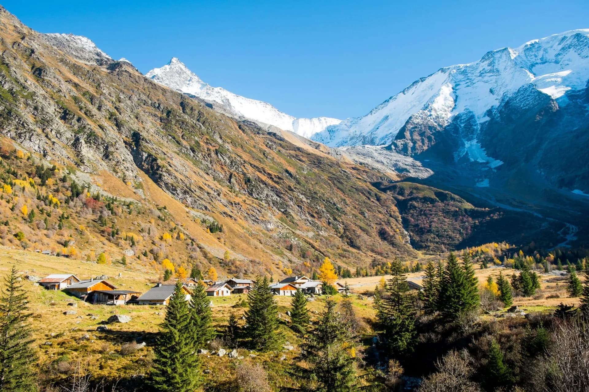 Alpine village nestled in valley with steep, snow-capped mountains and autumn foliage.