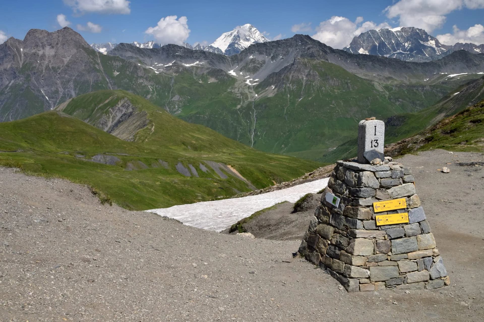 Stone trail marker on gravel path with green mountains and snow-capped peaks in background