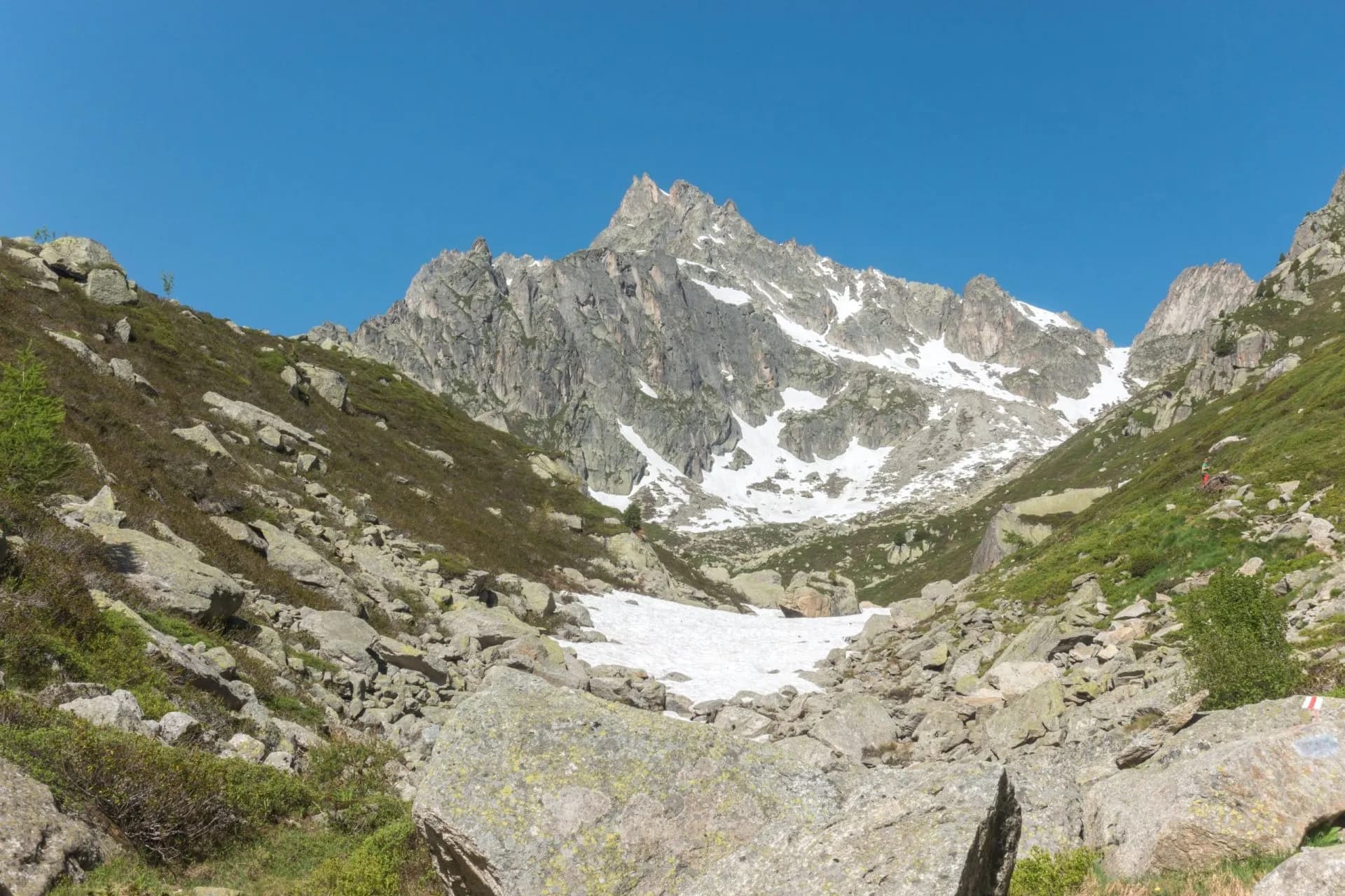 Rocky alpine valley with a snow patch leading to a jagged, snow-dusted mountain peak under a clear blue sky.