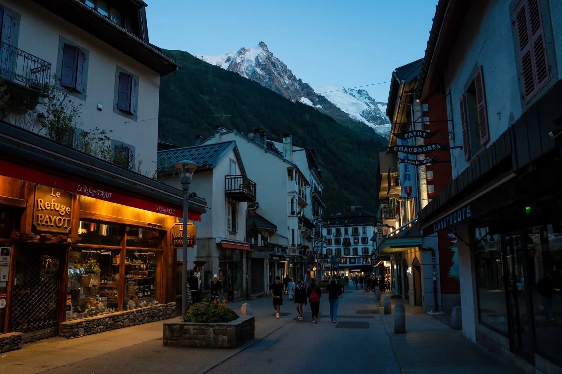 Pedestrians walking down a street toward a snow-capped mountain in Chamonix at dusk.