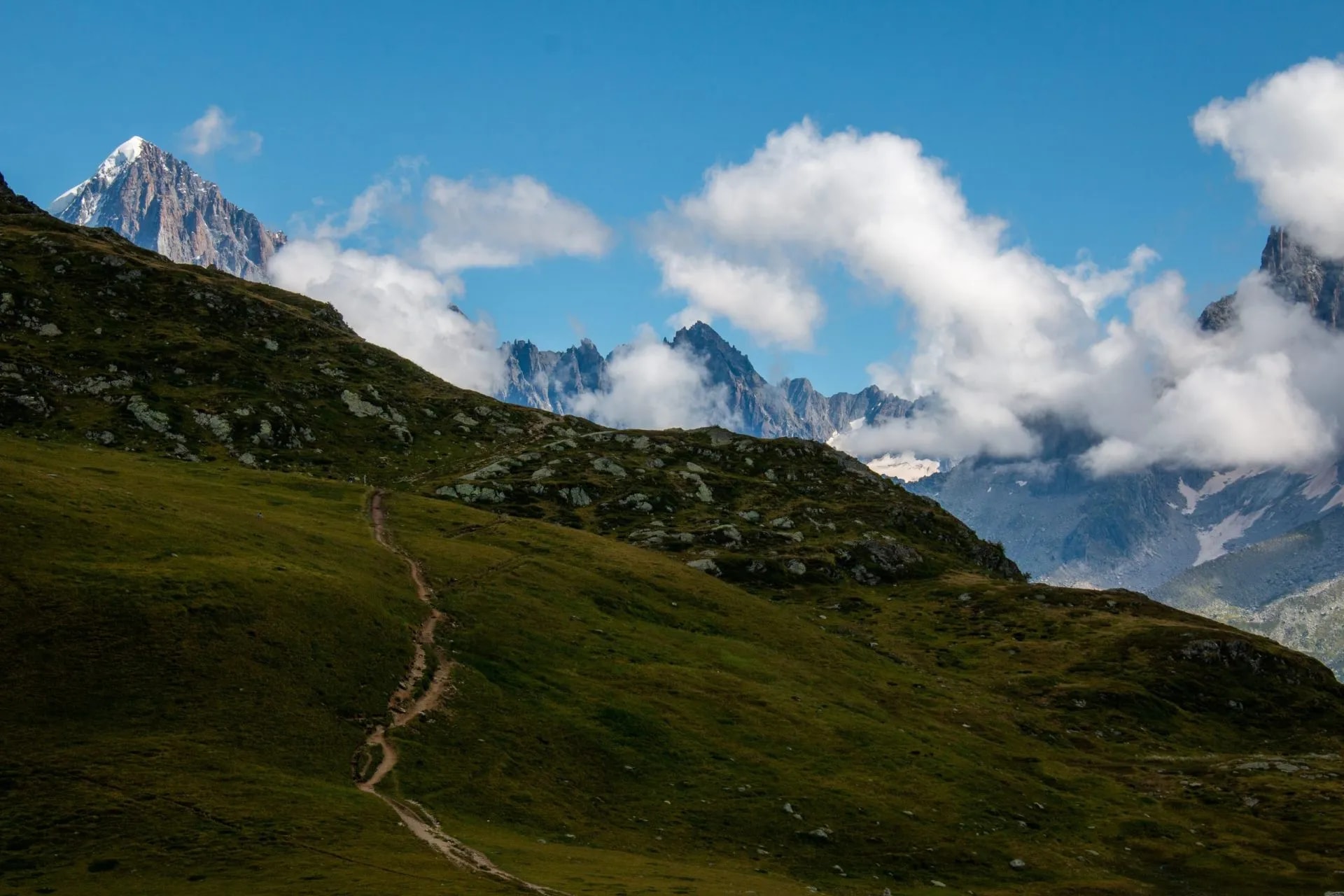 Hiking trail on grassy slope toward jagged, snow-capped mountains near Refuge de Bellachat.