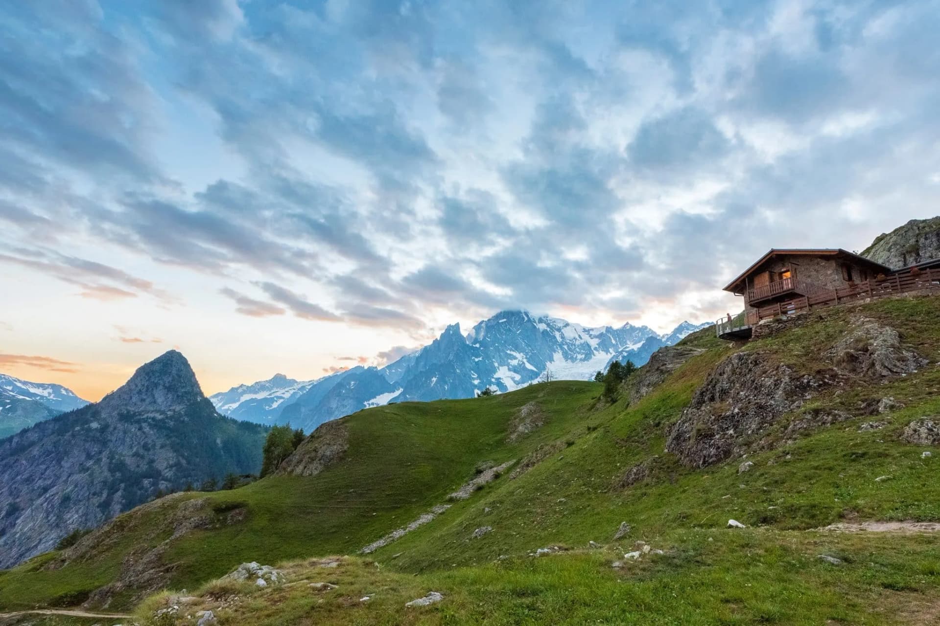 Mountain refuge Rifugio Bertone on grassy slope with snowy peaks under dramatic sky